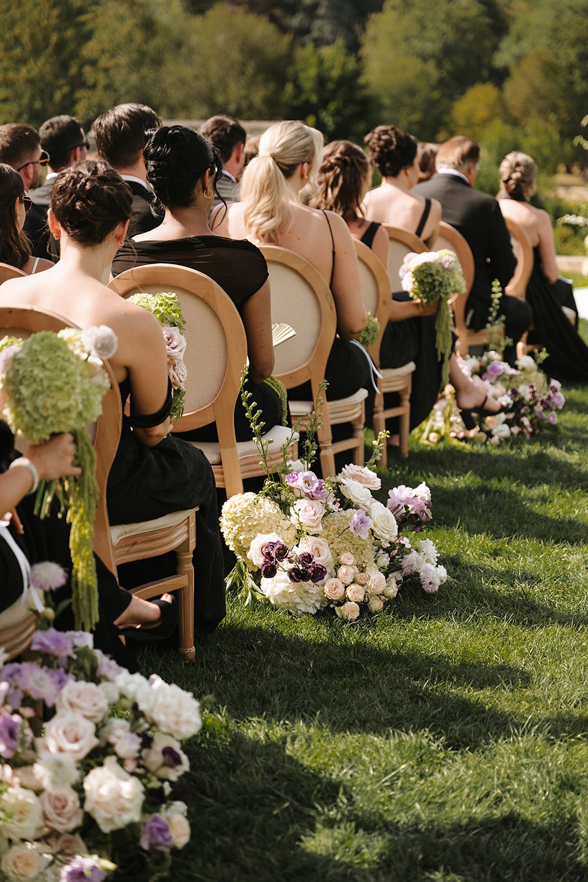 Guests in dark clothing sit on wooden chairs along a grass aisle edged with pastel flower arrangements.
