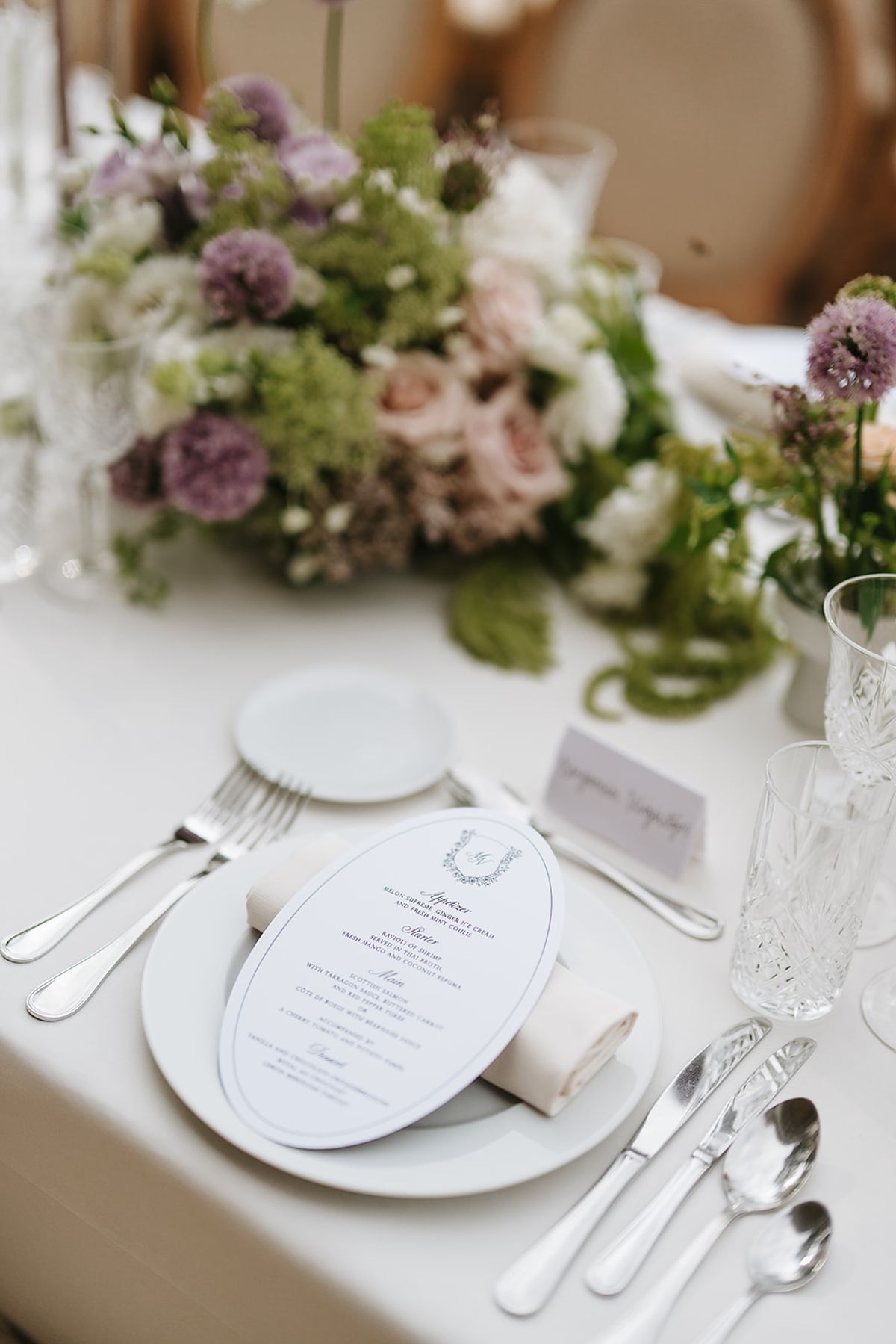 A dining table set with an oval menu card, cutlery, glassware and a centrepiece of lilac, pink and green flowers.