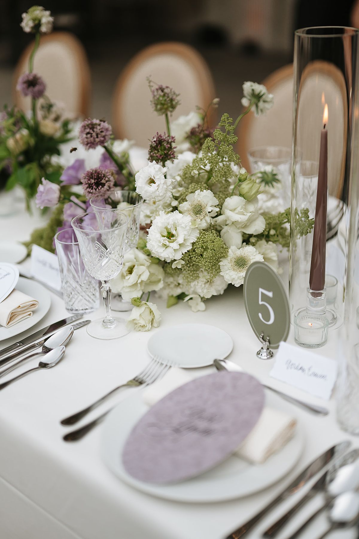 A white-clothed table set with crystal glasses, a lilac oval menu card, a taper candle, a table number and green and white flowers.