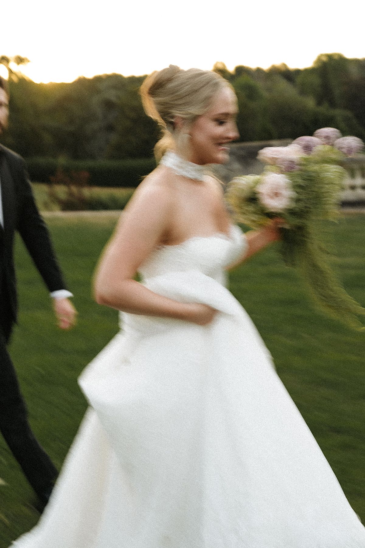 The smiling bride in a strapless white gown walks across a lawn holding a pink bouquet, a man in a dark suit beside her.