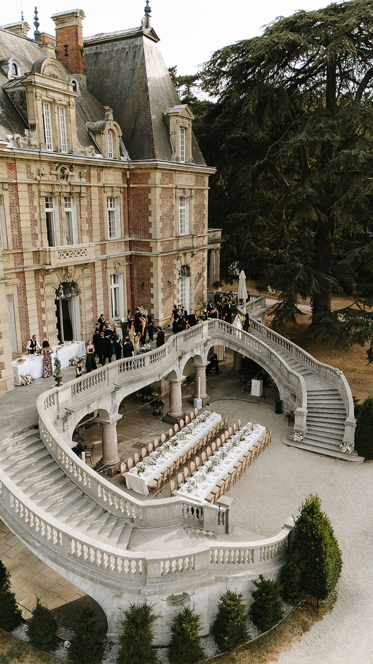 Seen from above, a brick chateau stands beside a curved stone staircase with a long dining table laid out on the terrace below.