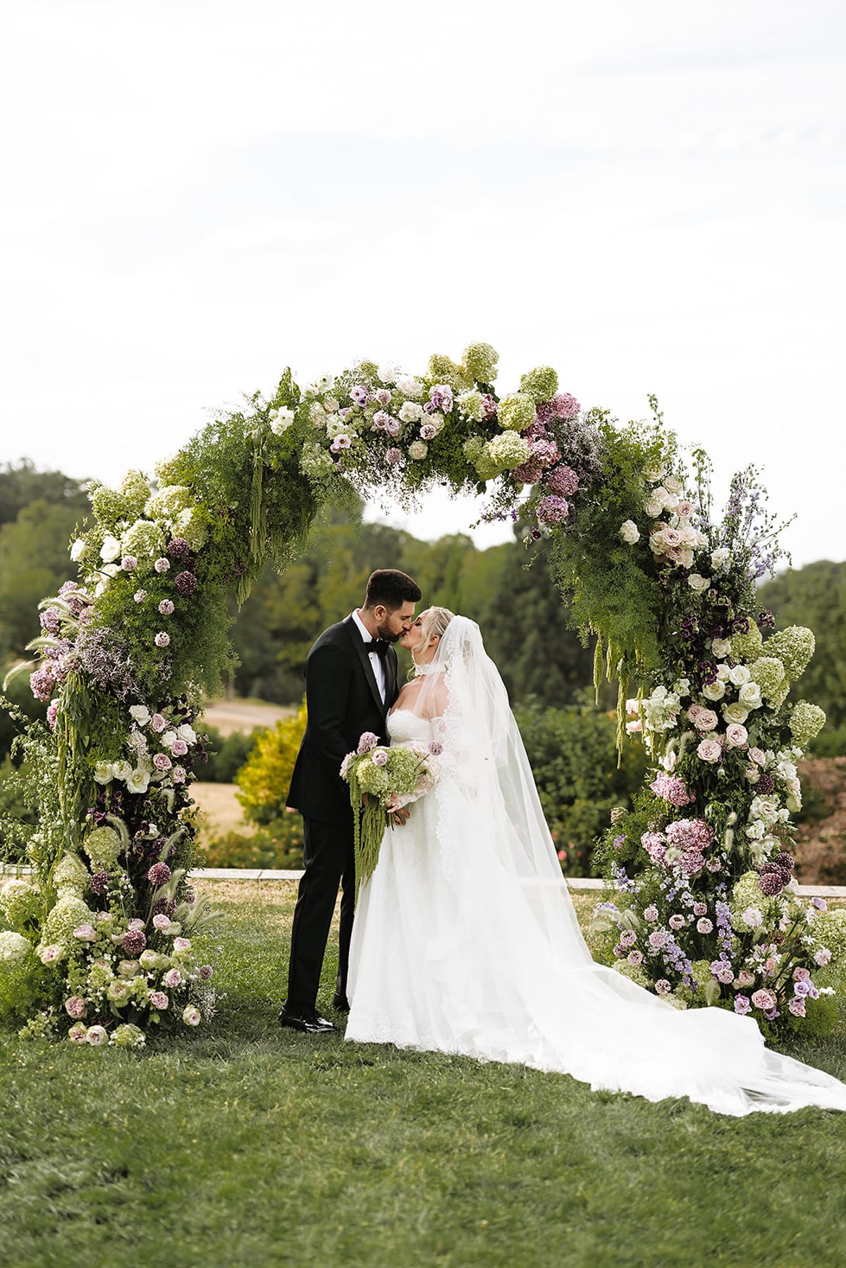 The bride and groom kiss in front of a large round arch of lilac, white and green flowers on a lawn.