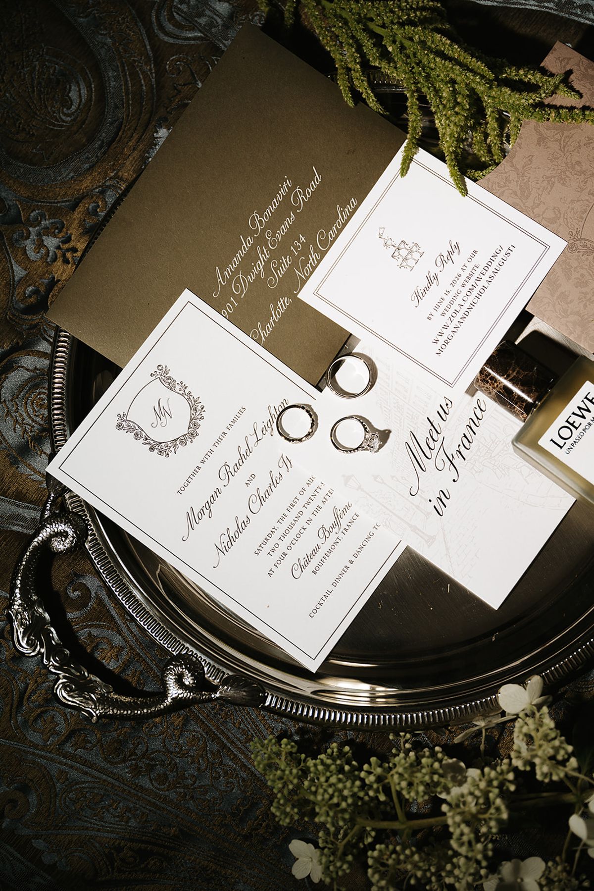 Letterpress invitation cards and two wedding rings rest on an ornate silver tray with sprigs of green foliage.