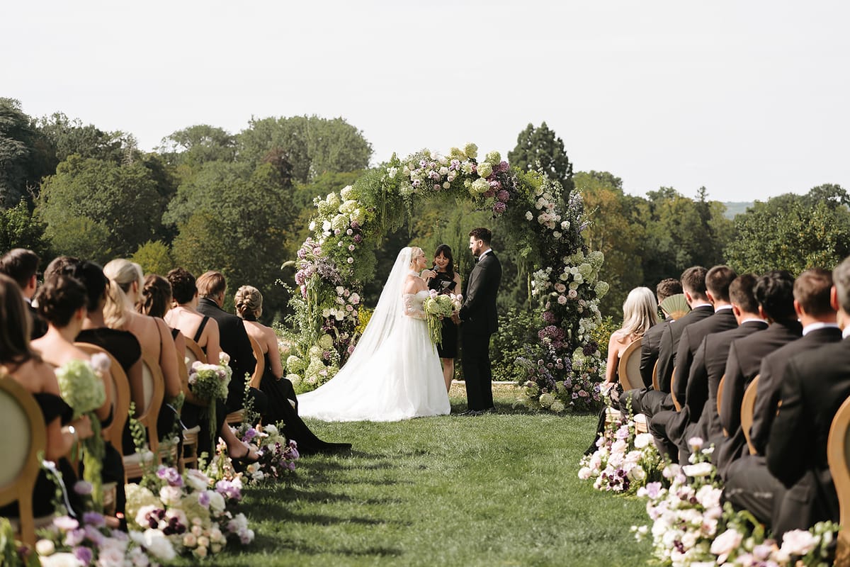 Guests sit on either side of a grass aisle lined with flowers as the couple stand beneath a floral arch.