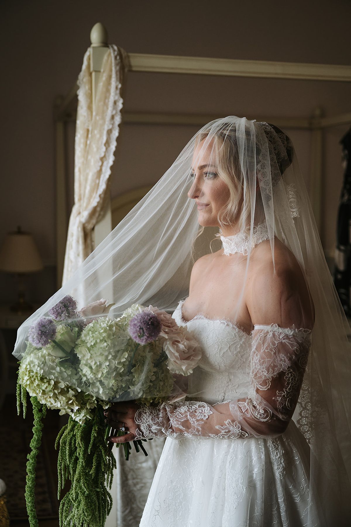 The bride, wearing a sheer veil and lace off-shoulder sleeves, looks aside while holding a bouquet of pale green hydrangeas.