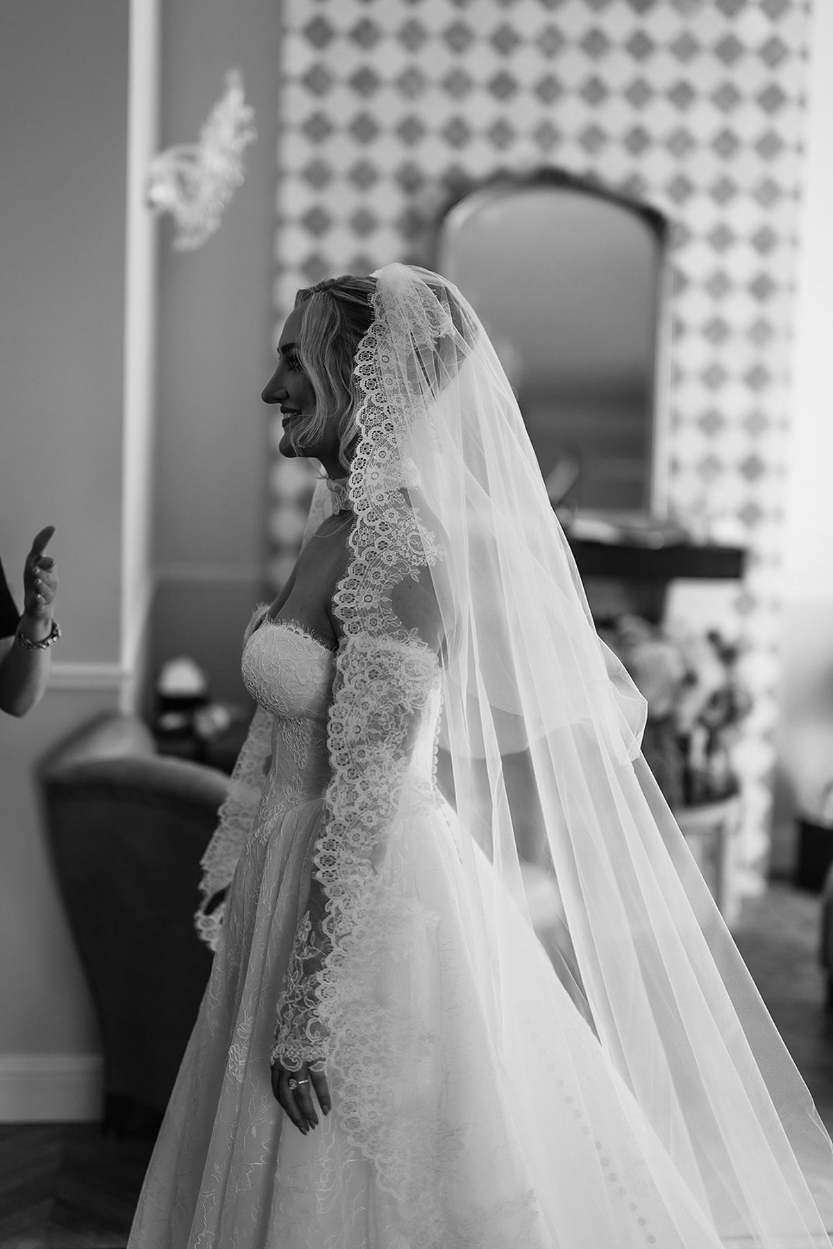 The bride, wearing a long veil and lace sleeves, is seen in profile in a room with patterned wallpaper.