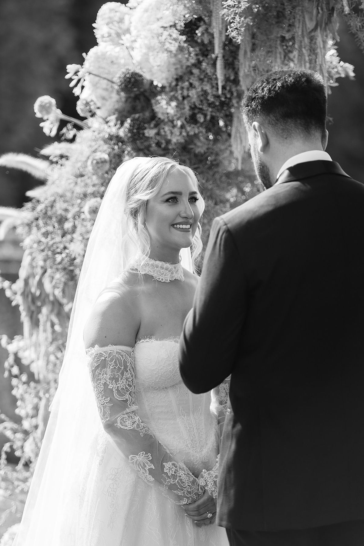 The bride in a veil and lace gloves smiles at the groom, who stands with his back to the camera, among flowers.