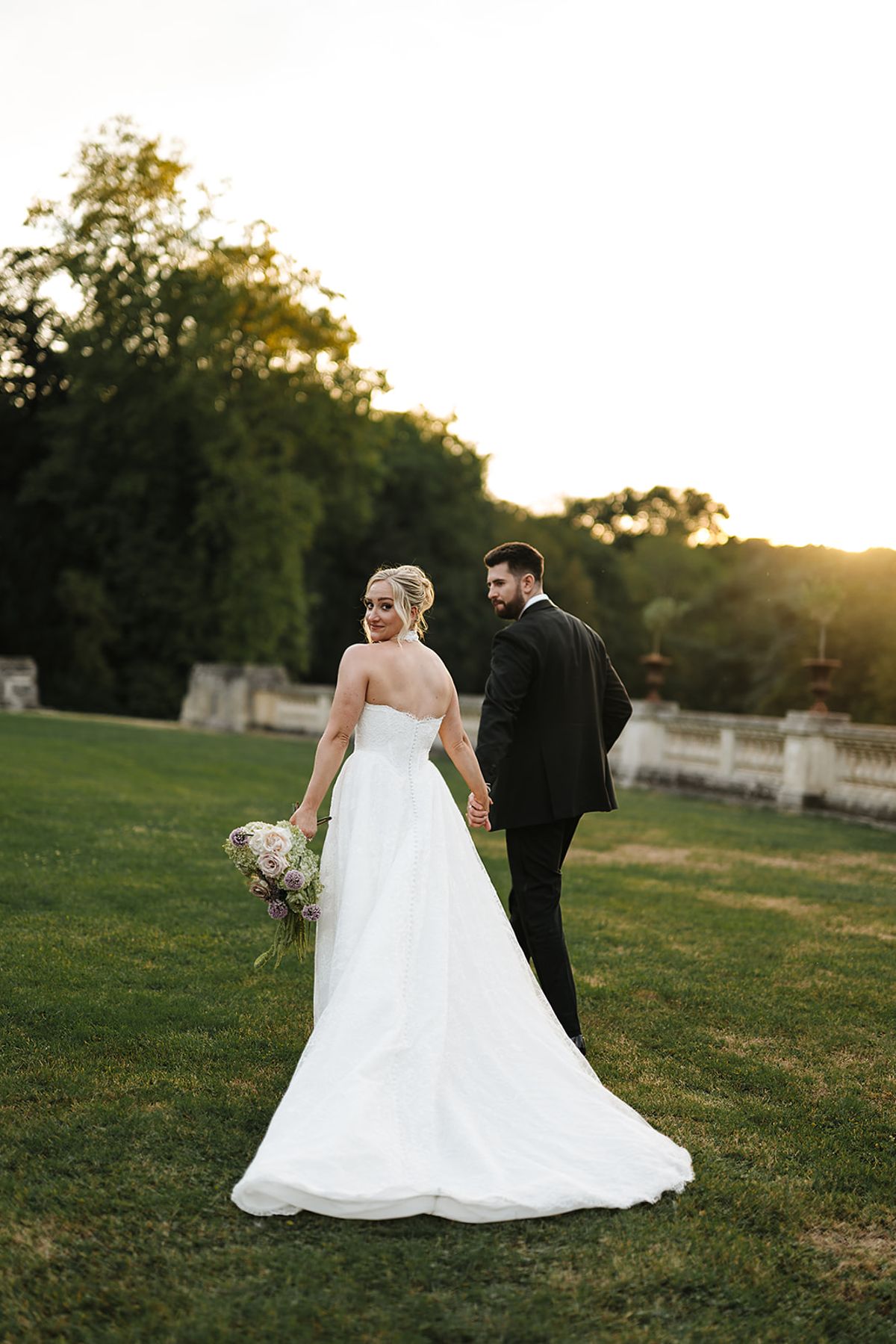 The bride in a long white gown and the groom in a dark suit walk away across a grassy lawn in warm low light.