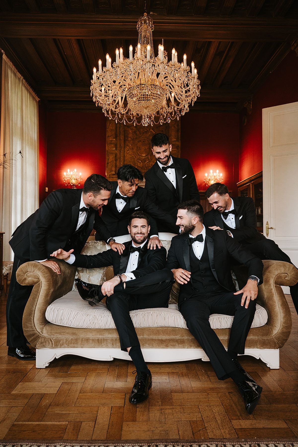 Six men in black tuxedos sit and lean together on a cream sofa in a room with red walls and a gold chandelier.