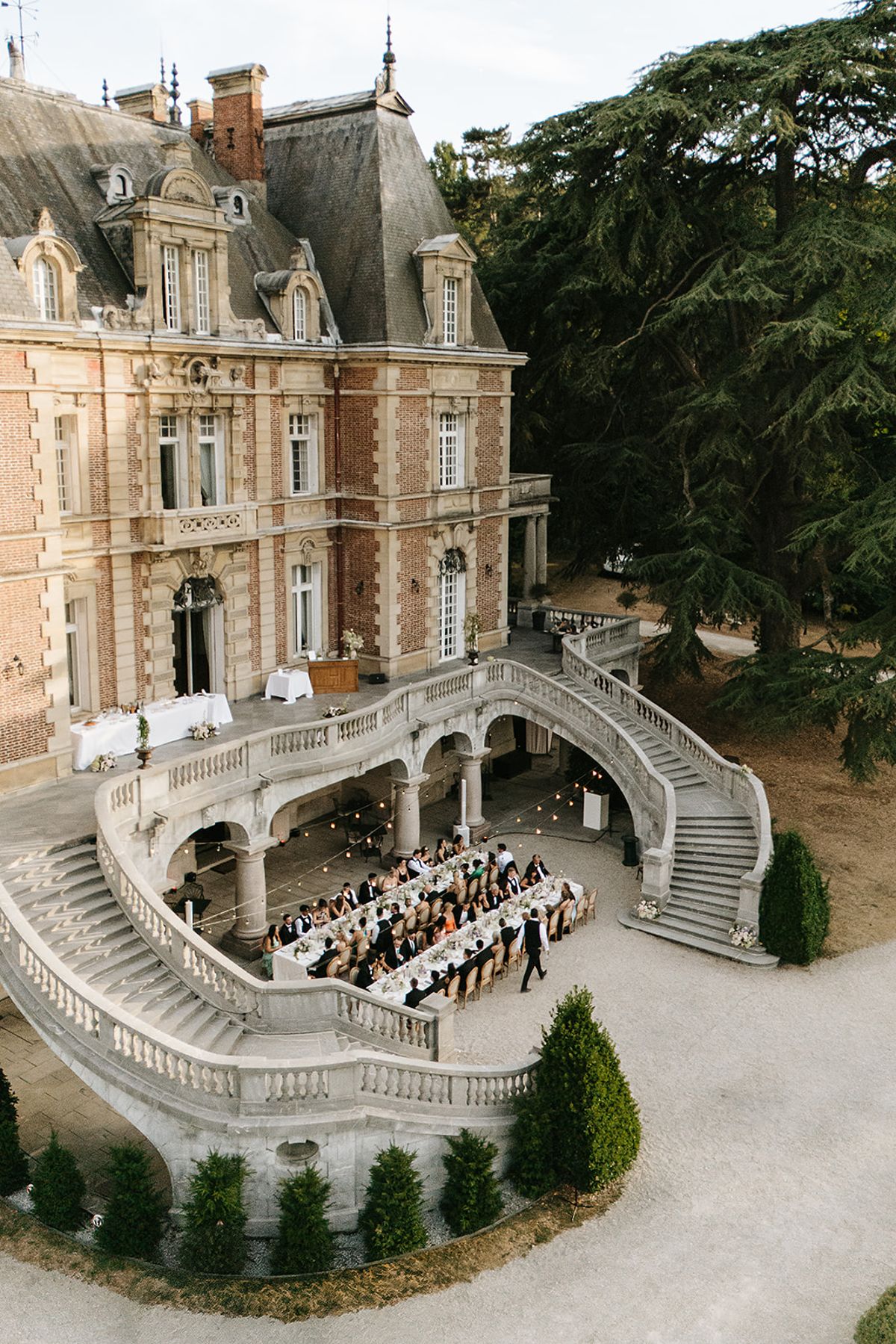 A drone view of a long dinner table with guests beneath a curved stone staircase at the front of a brick chateau.