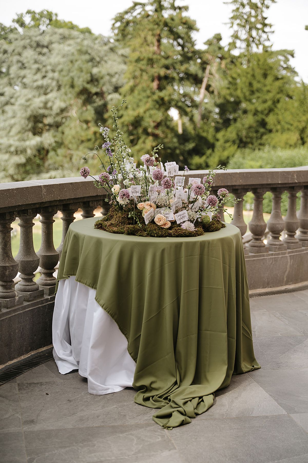 A round table draped in an olive green cloth holds a low arrangement of pink and cream flowers beside a stone balustrade, with trees beyond.