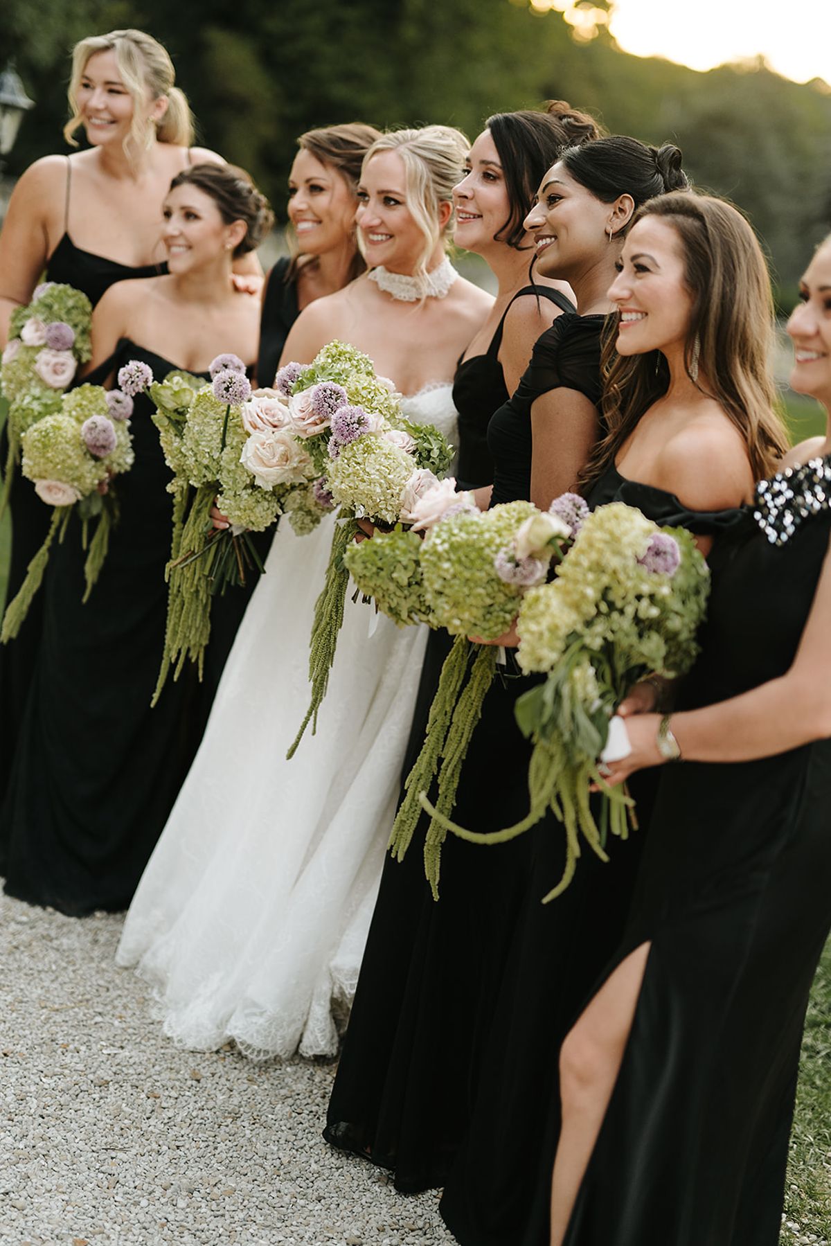 The bride in a white gown and bridesmaids in black dresses laugh together, holding large green and pale bouquets in warm light.