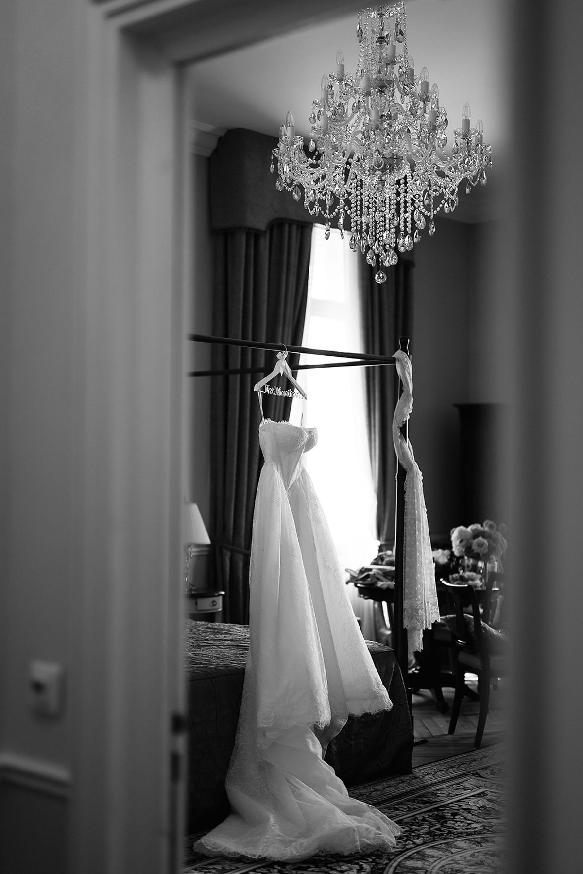 Seen through a doorway in black and white, a white wedding dress hangs from a rail beneath a crystal chandelier in an elegant bedroom.