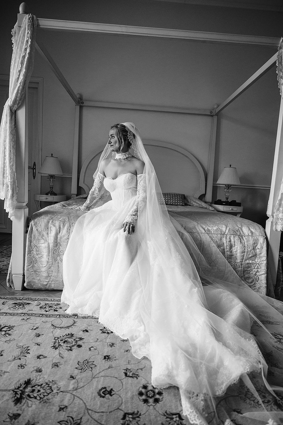 In black and white, the bride in a full tulle gown and long veil sits on the edge of a canopy bed.