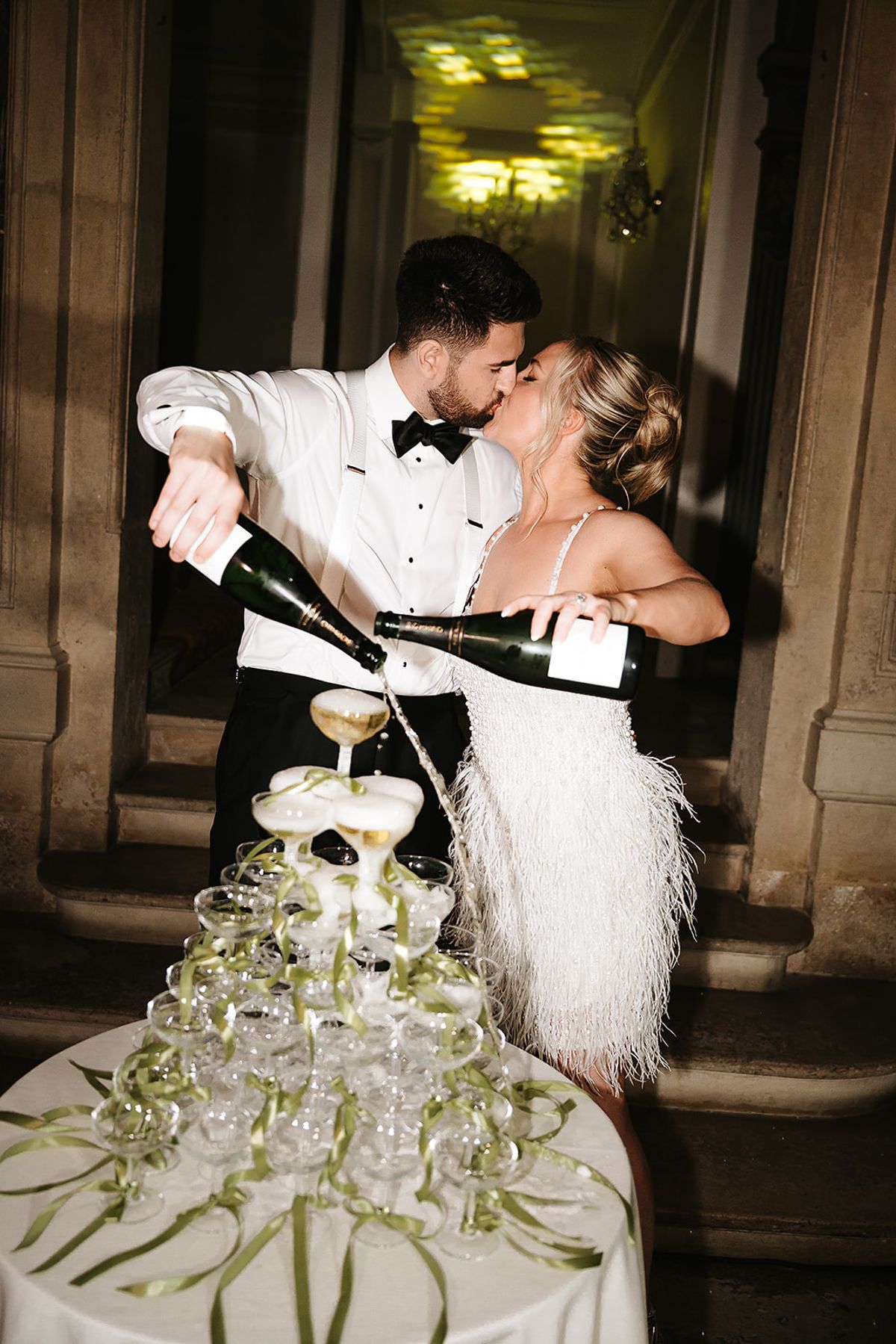 The couple kiss as they both pour champagne from bottles into a tall tower of glasses on a table decorated with ribbons.