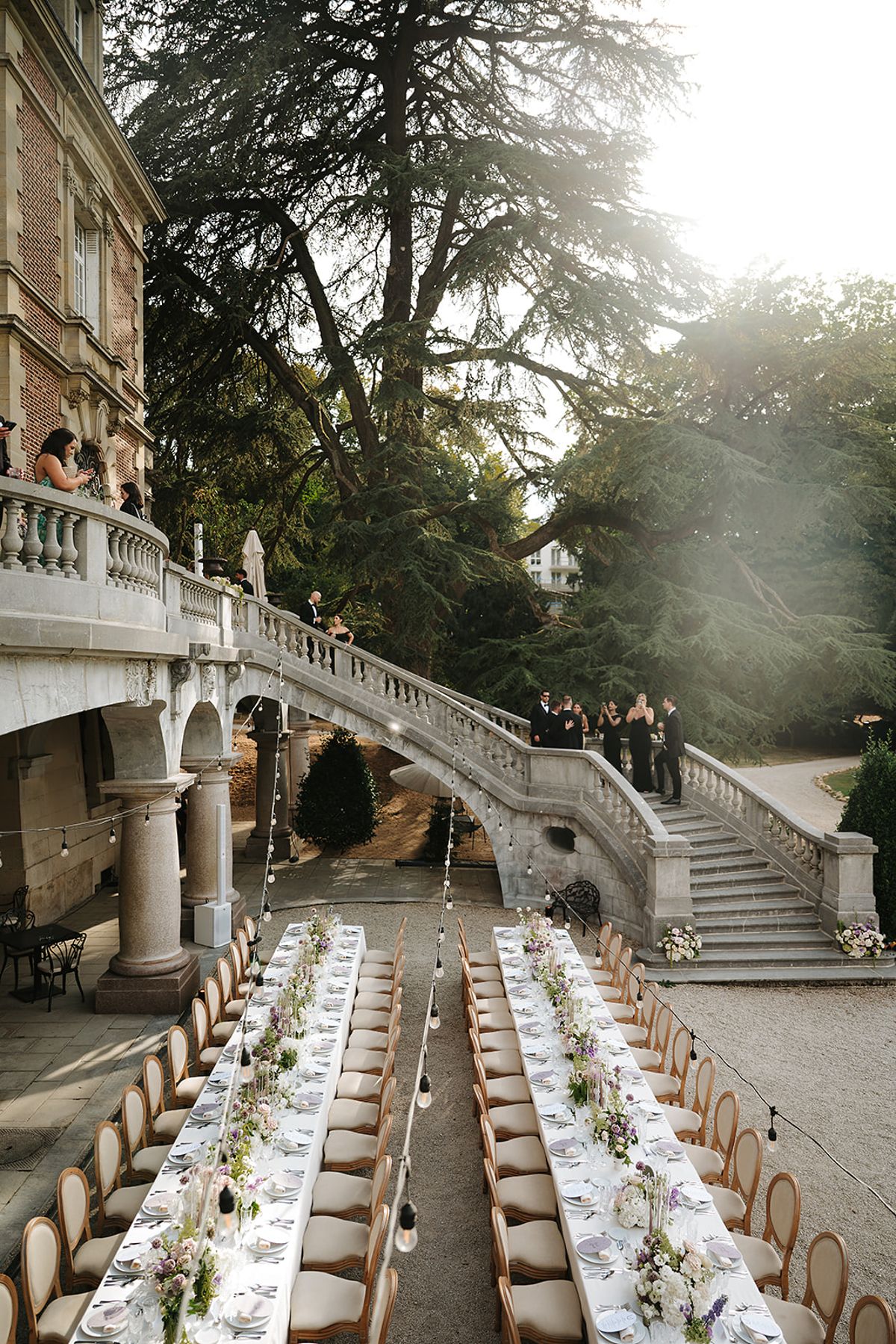 Guests in dark clothes stand on a stone staircase beside two long, flower-decked dining tables set outdoors under tall trees.