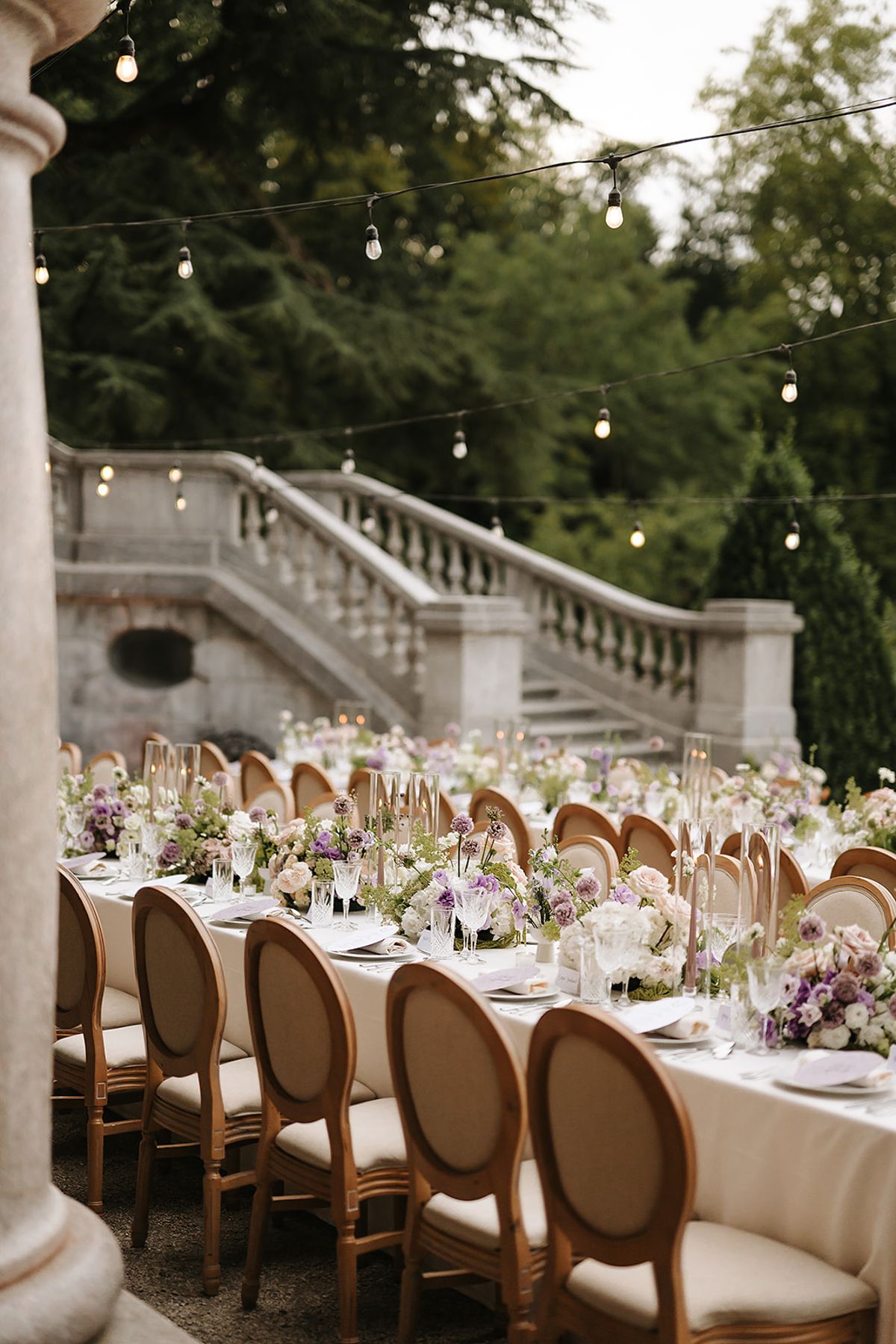 A long dining table dressed with pale flowers and wooden chairs runs along a grey stone staircase framed by tall trees.