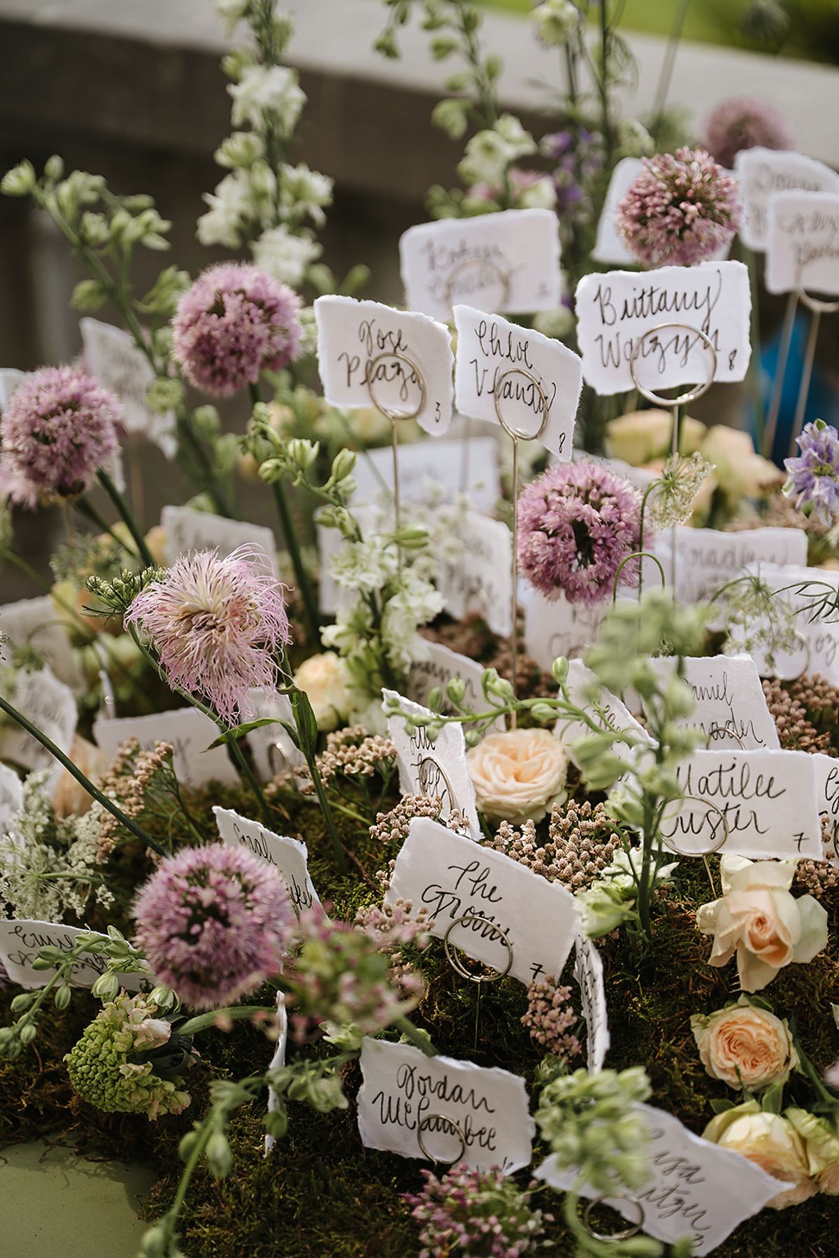 Small white cards with handwritten names are tucked into an arrangement of lilac alliums, cream roses and green foliage.