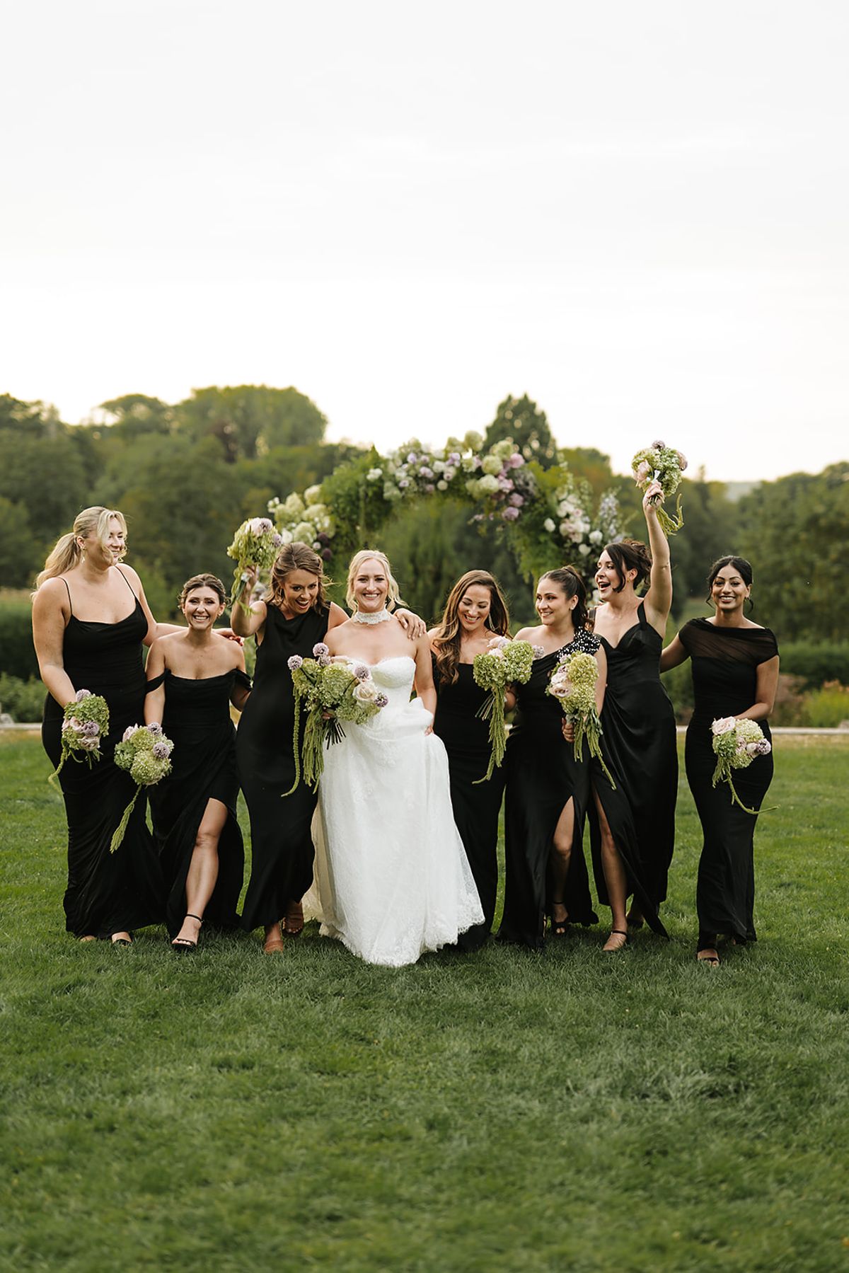 The bride in a white gown walks towards the camera among smiling bridesmaids in black dresses, with a floral arch behind them.