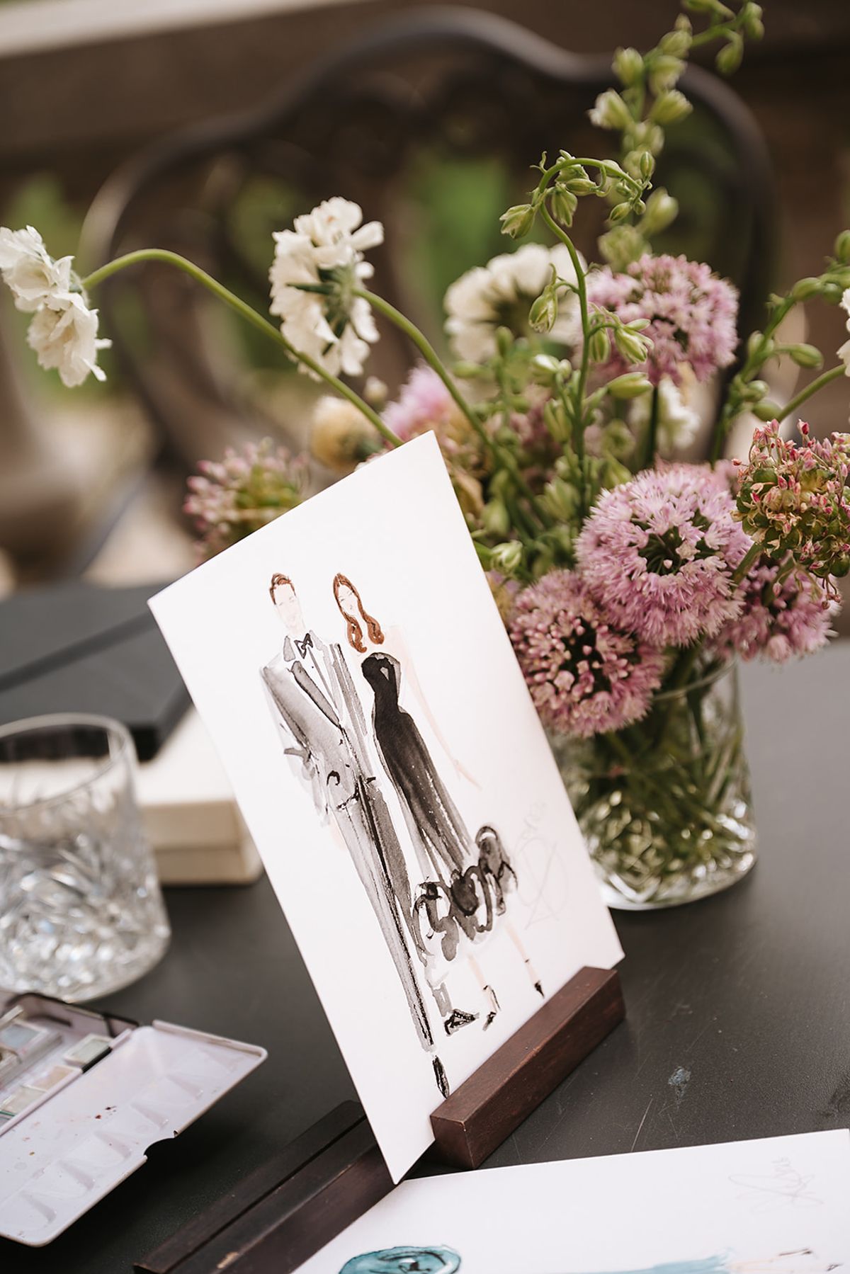 A framed watercolour sketch of figures stands on a dark table beside a glass vase of pink and white flowers.