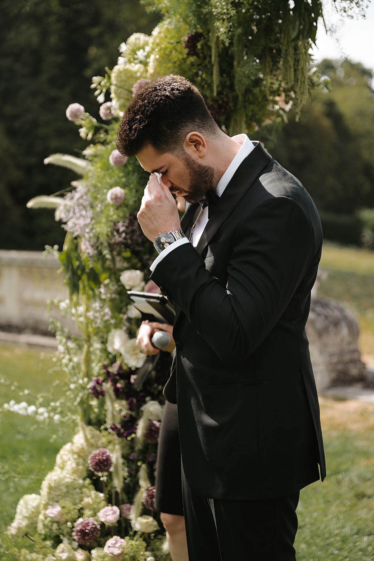 The groom, in a black tuxedo, wipes away a tear beside a tall arch of white and plum flowers.