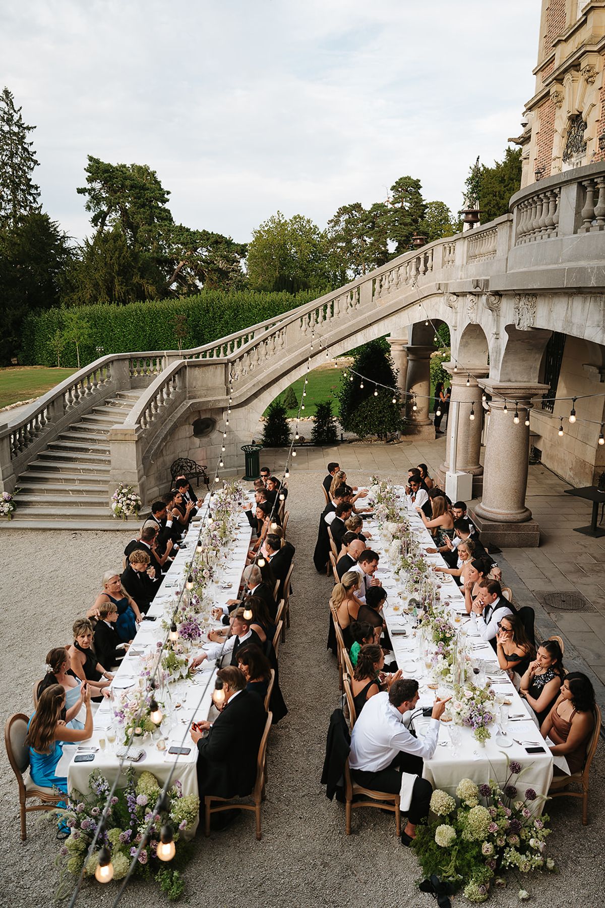 Seen from above, guests sit along two long tables decorated with flowers and candles beside a sweeping stone staircase and lawn.