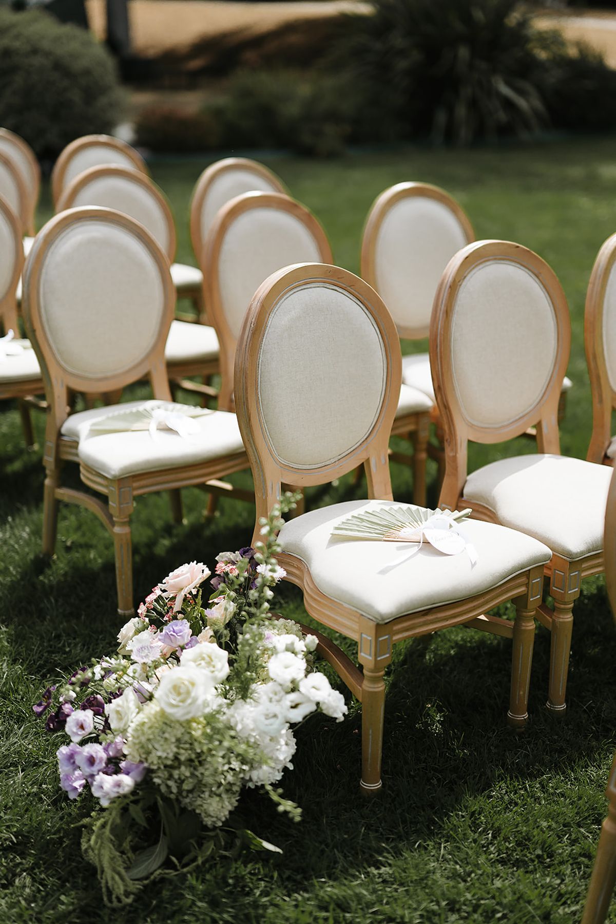 Gold-framed round-backed chairs with cream cushions stand in rows on grass, with a white and lilac floral arrangement at the aisle.