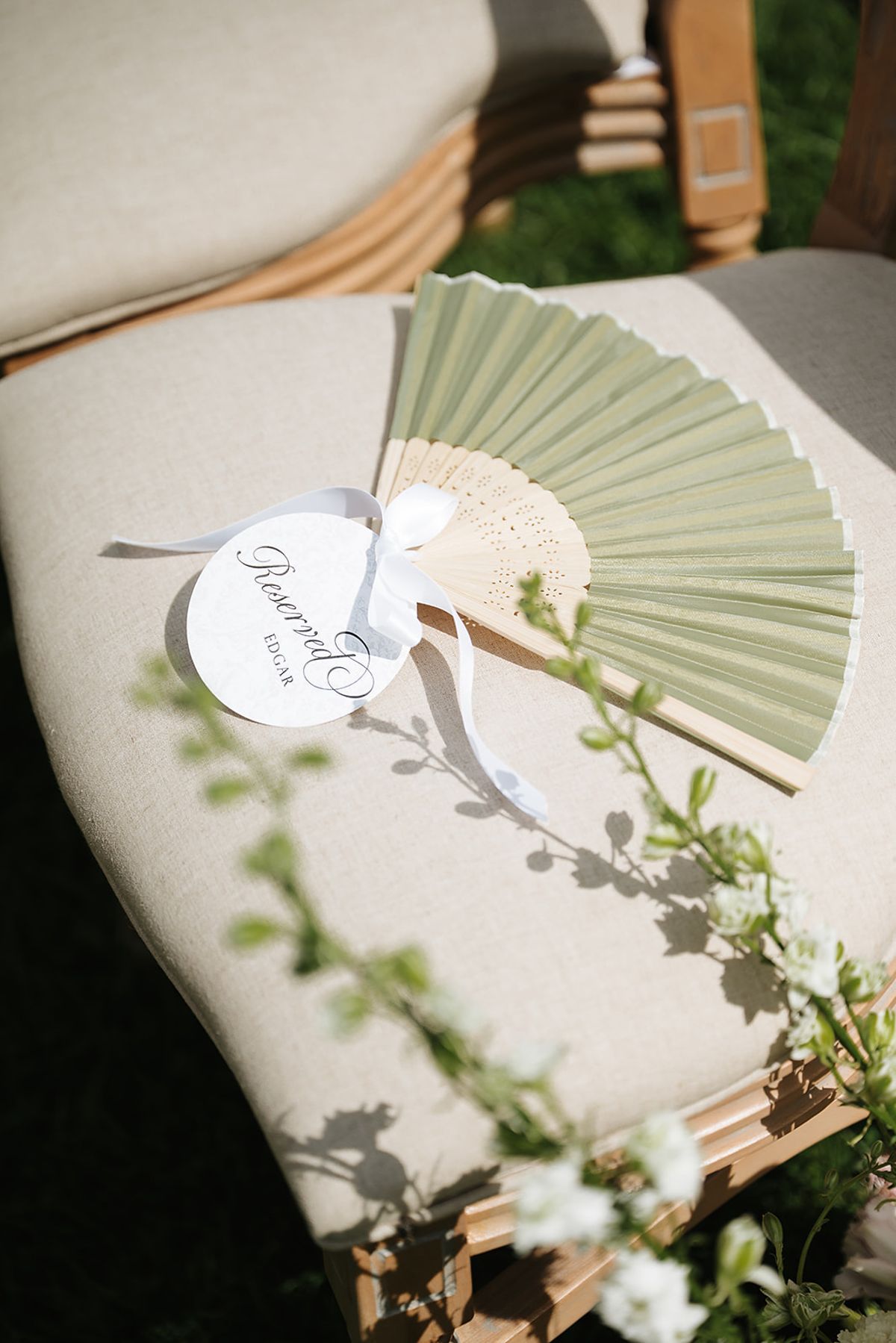 A sage green paper fan with a small white tag lies on a cream upholstered chair trimmed with trailing foliage.