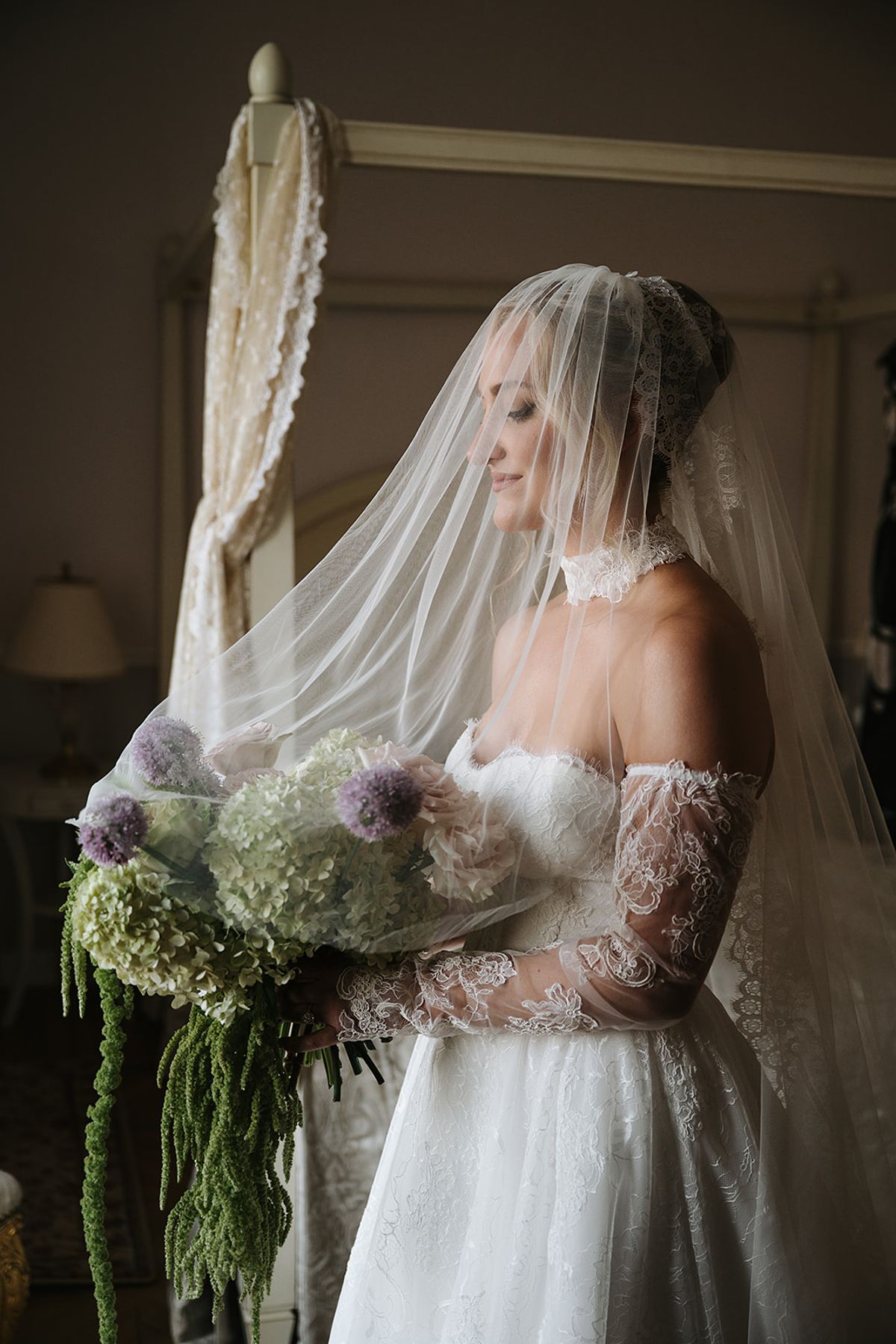 The bride in a lace-sleeved gown and sheer veil stands in profile in a dim room, holding a large lilac and green bouquet.