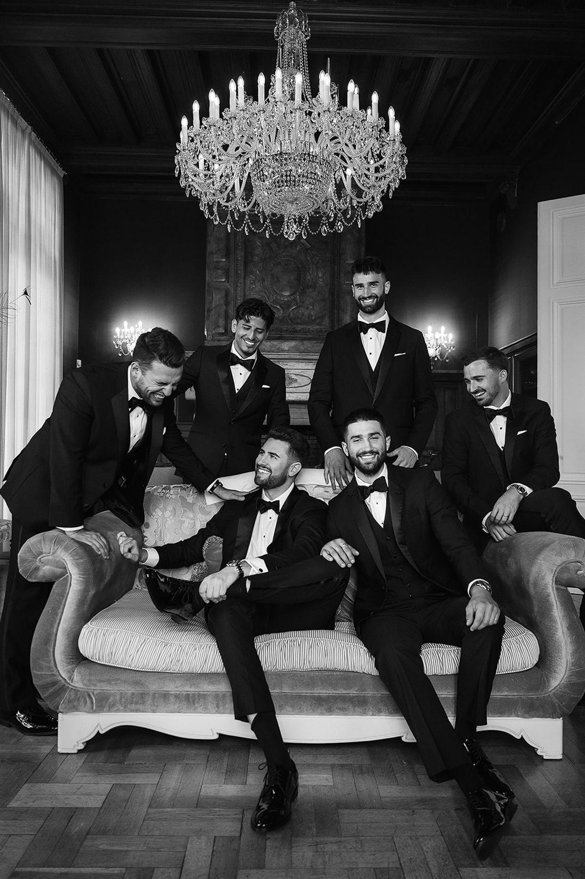 In black and white, seven men in tuxedos sit and lean around a sofa beneath a large crystal chandelier.