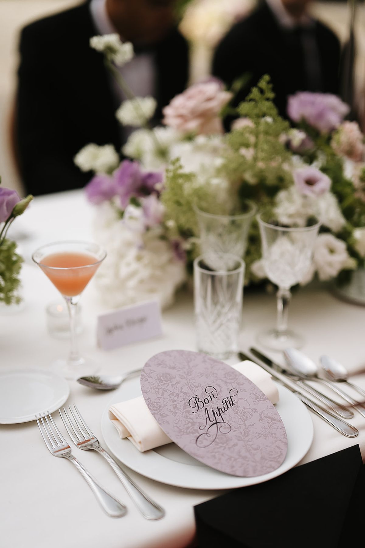 A place setting with a lilac oval menu card with a French greeting in script on a folded napkin, crystal glasses, a peach cocktail and a centrepiece of white and lilac flowers, with guests in dark suits blurred behind.
