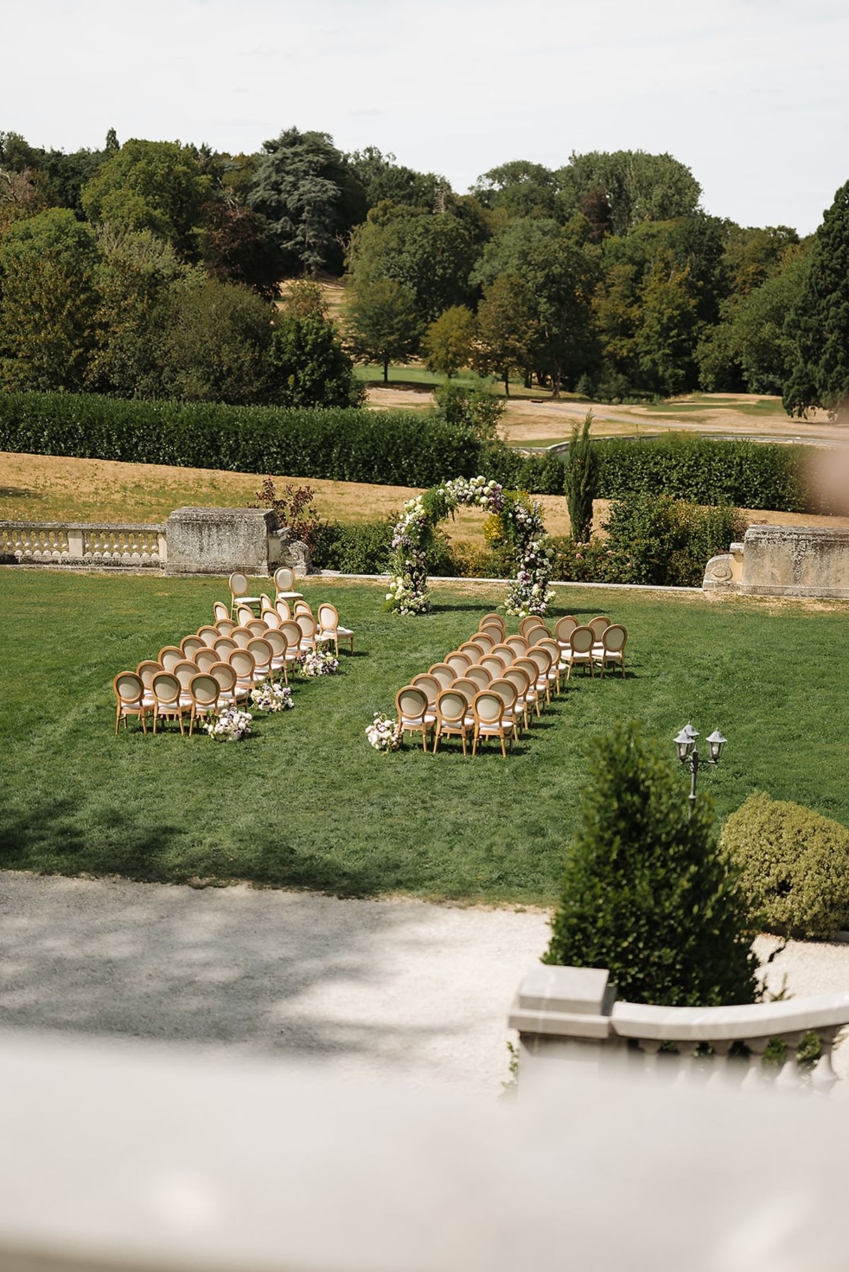 Rows of wooden chairs and a floral arch stand on a green lawn, with wooded parkland and hedges in the distance.