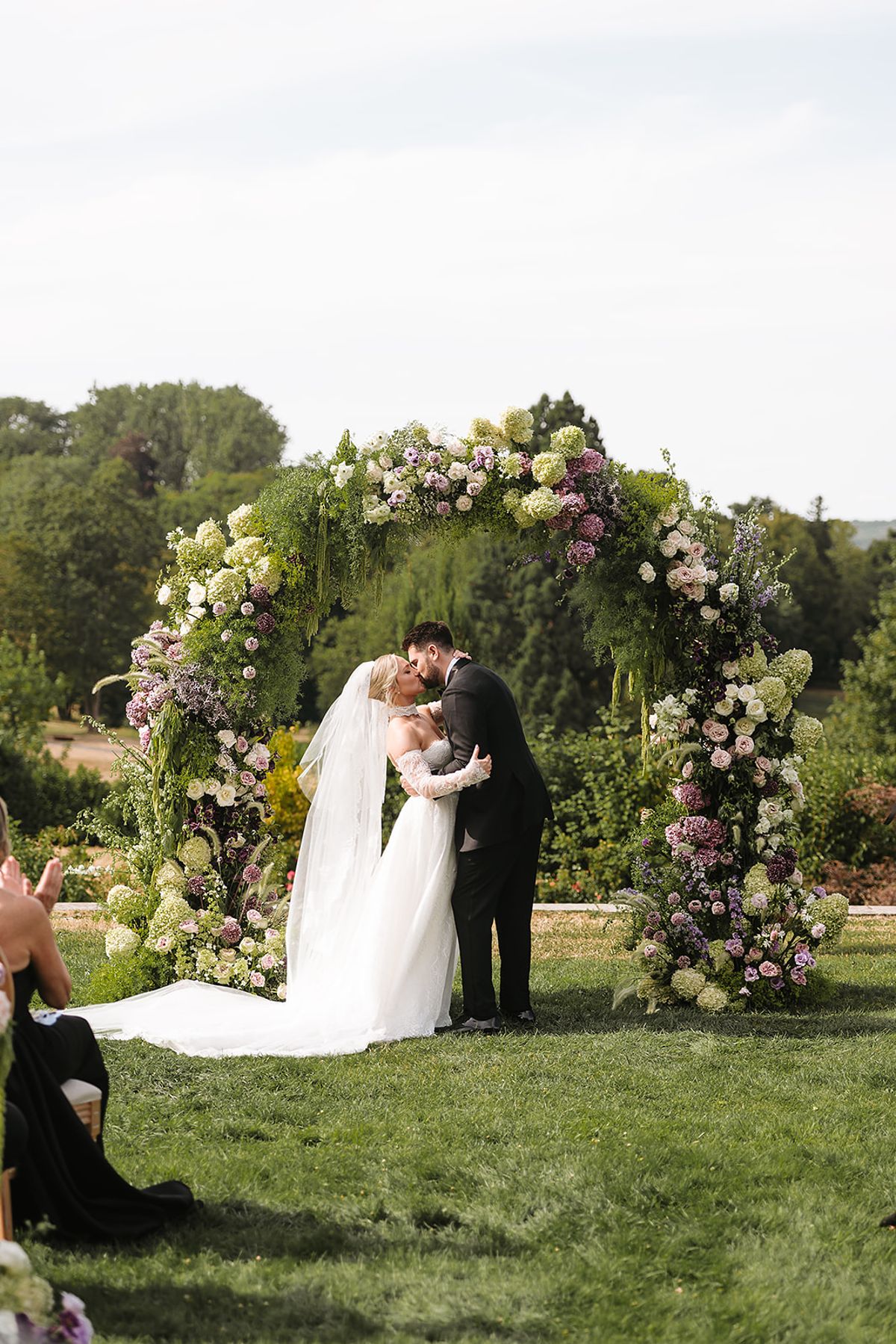 The bride and groom kiss beneath a floral arch of white, green and purple blooms on a lawn, as a guest watches nearby.