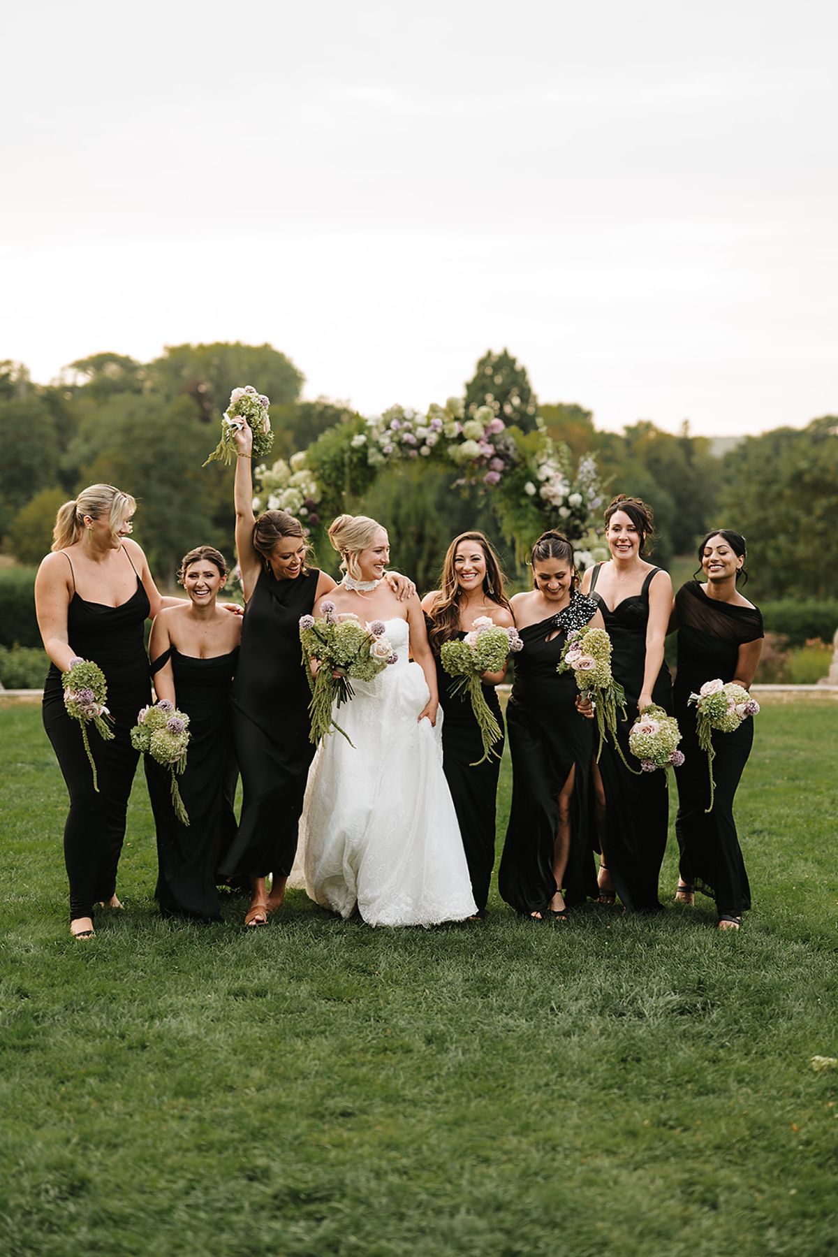 The bride and her bridesmaids in black dresses walk across a lawn, smiling and raising their bouquets.