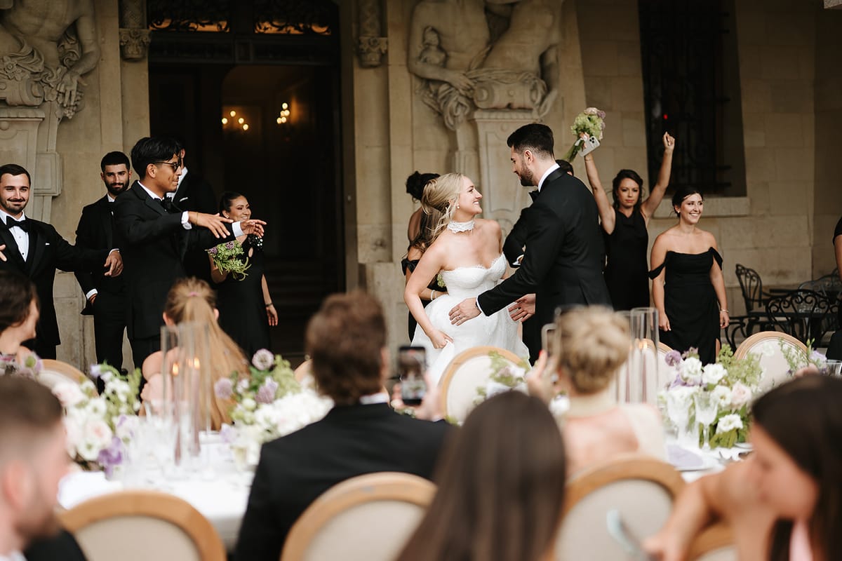 The bride, groom and dark-dressed wedding party cheer and raise their arms in front of a stone château while seated guests look on.