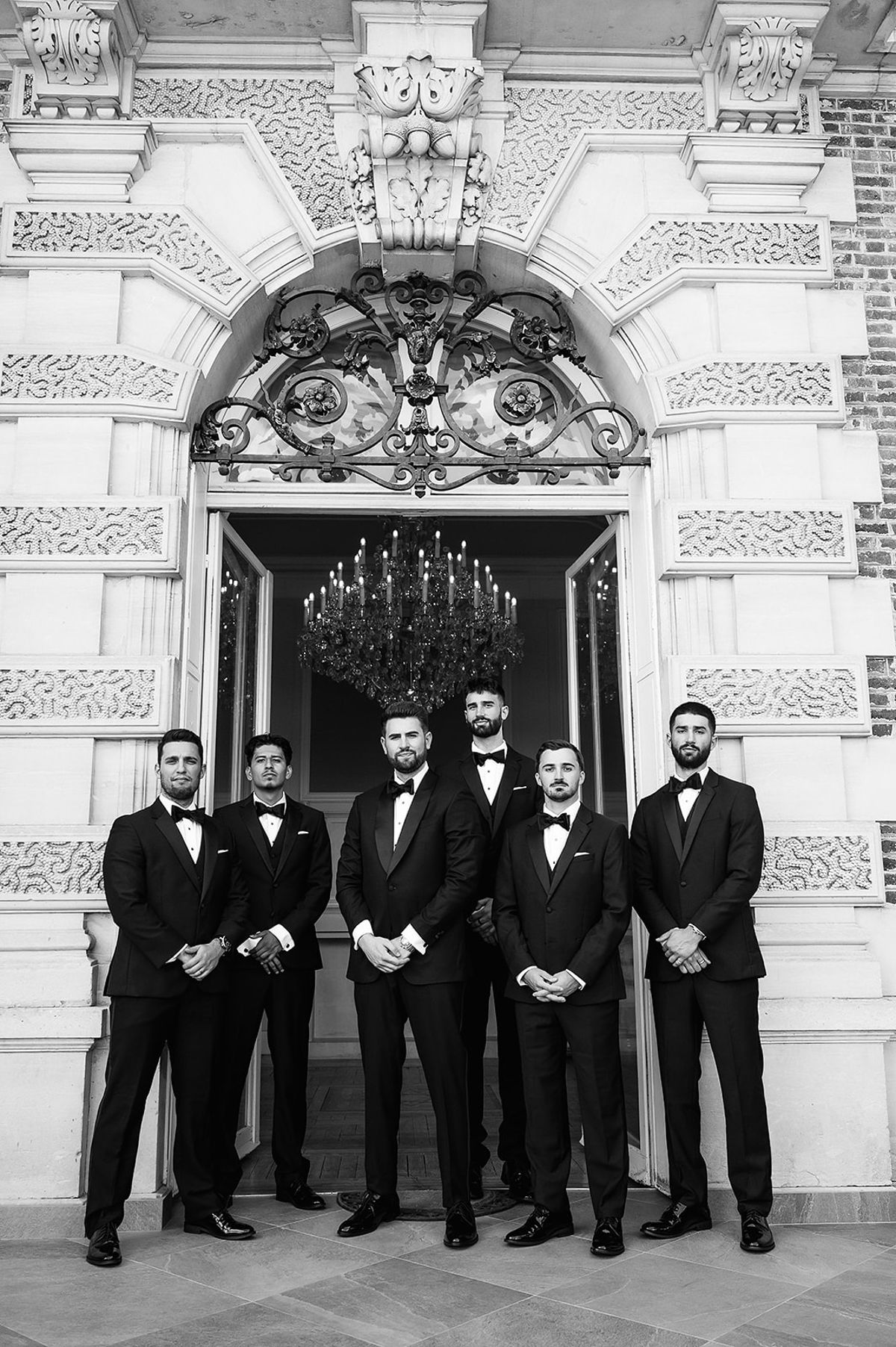 Six men in dark tuxedos stand in a row in front of an ornate arched chateau doorway with a chandelier inside.