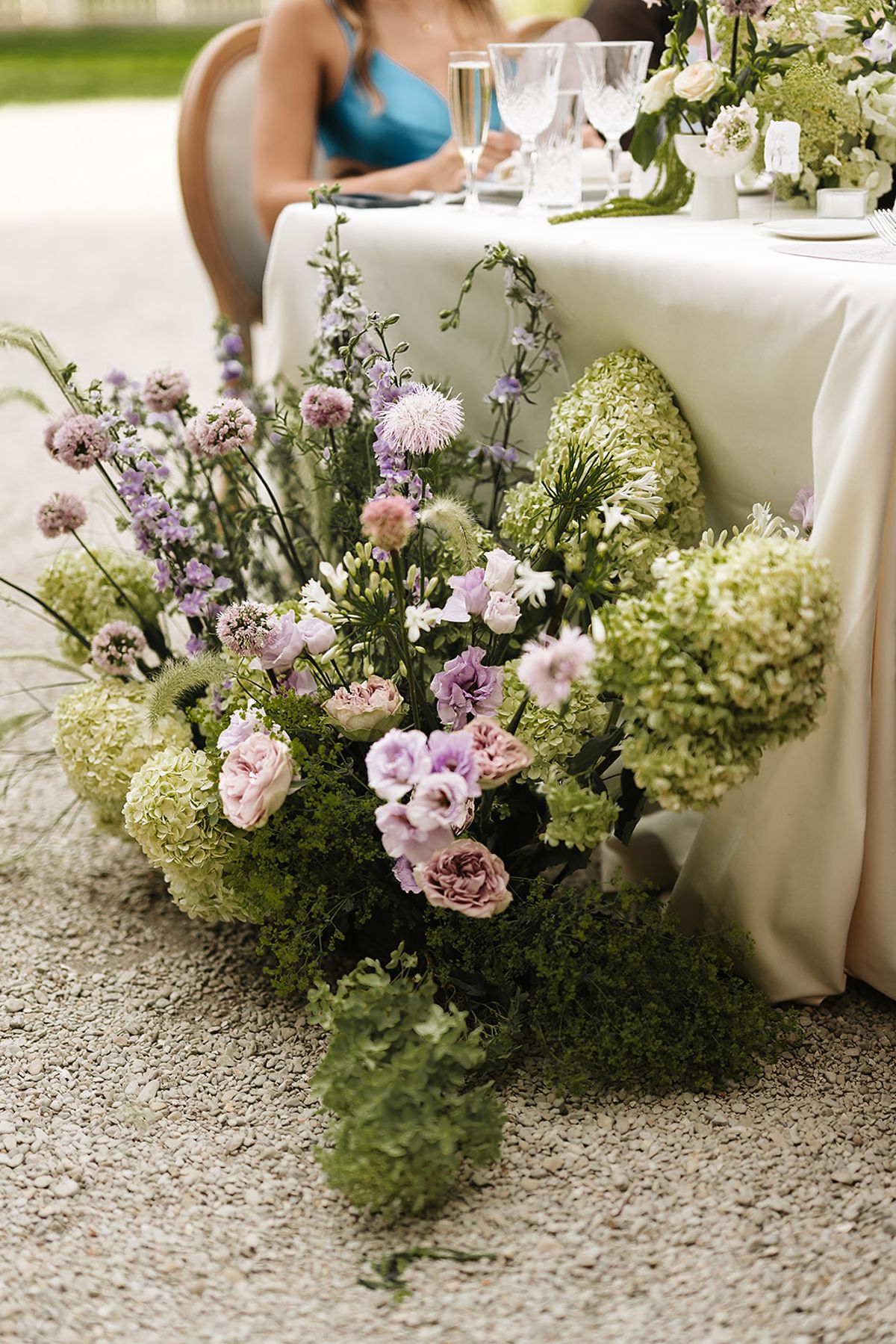 A low arrangement of green hydrangeas, lilac lisianthus, pink roses and delphiniums spills from the foot of a white-clothed table onto the gravel, with a guest seated behind it.