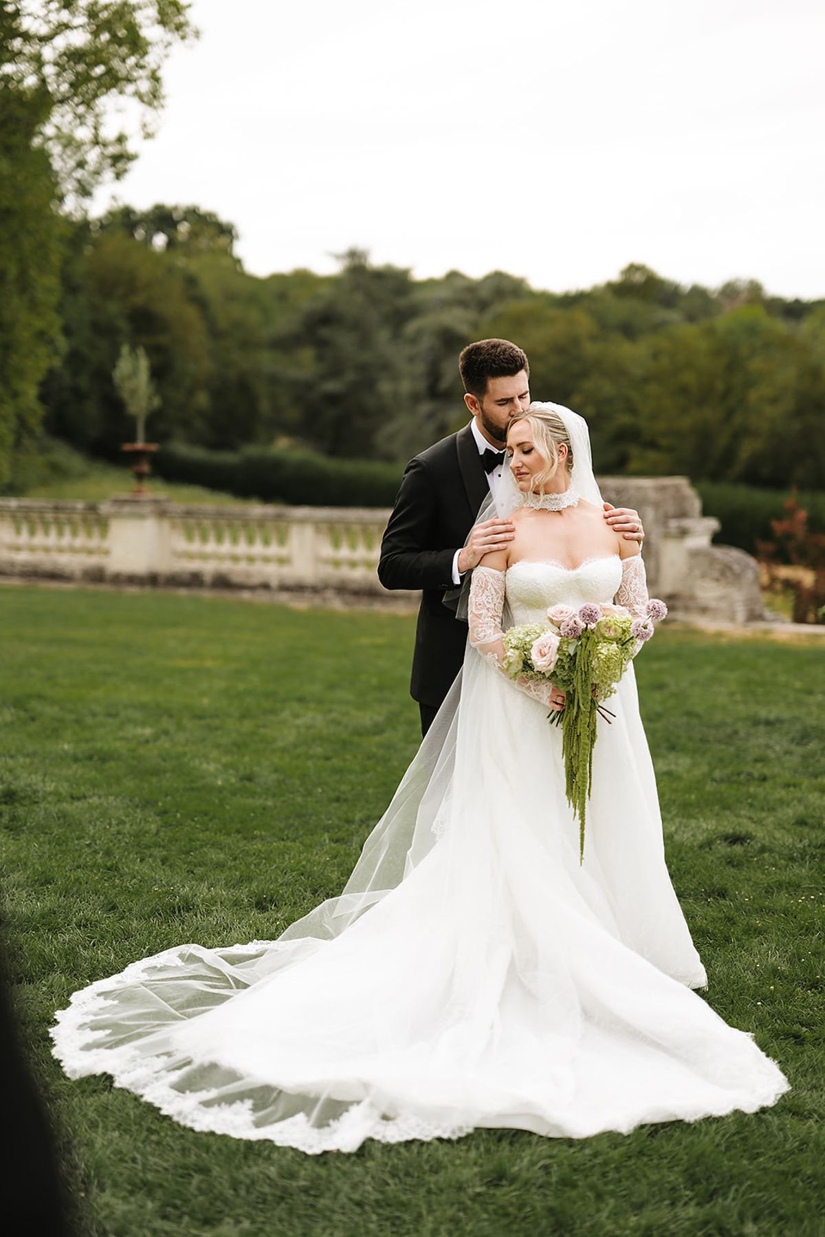 The groom stands behind the bride and holds her close on a lawn, her gown and long train spread across the grass.