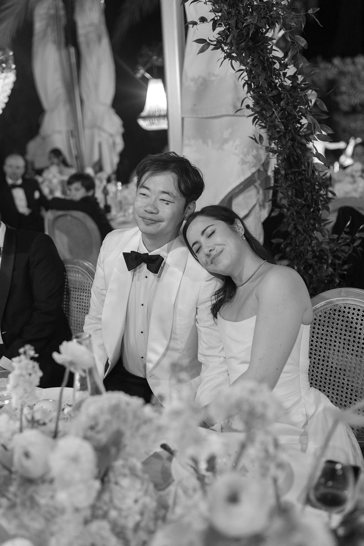 Black and white view of Carina resting her head on Stefan's shoulder at the dinner table
