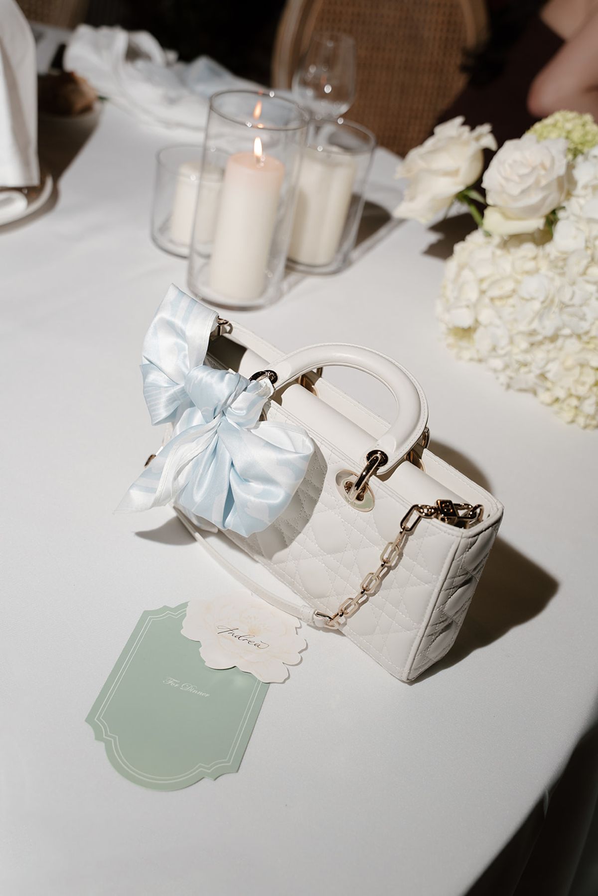 A white handbag tied with a pale blue bow on a dinner table beside candles