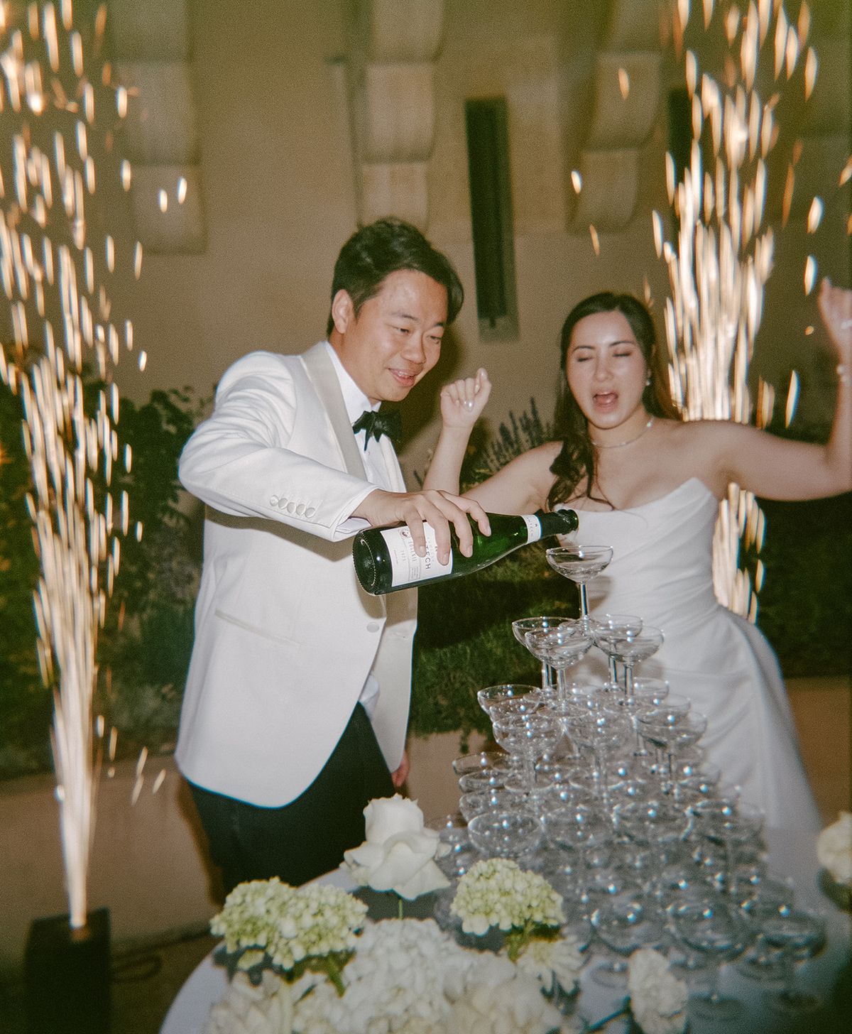 Stefan pouring champagne into the tower as Carina cheers, sparklers behind them