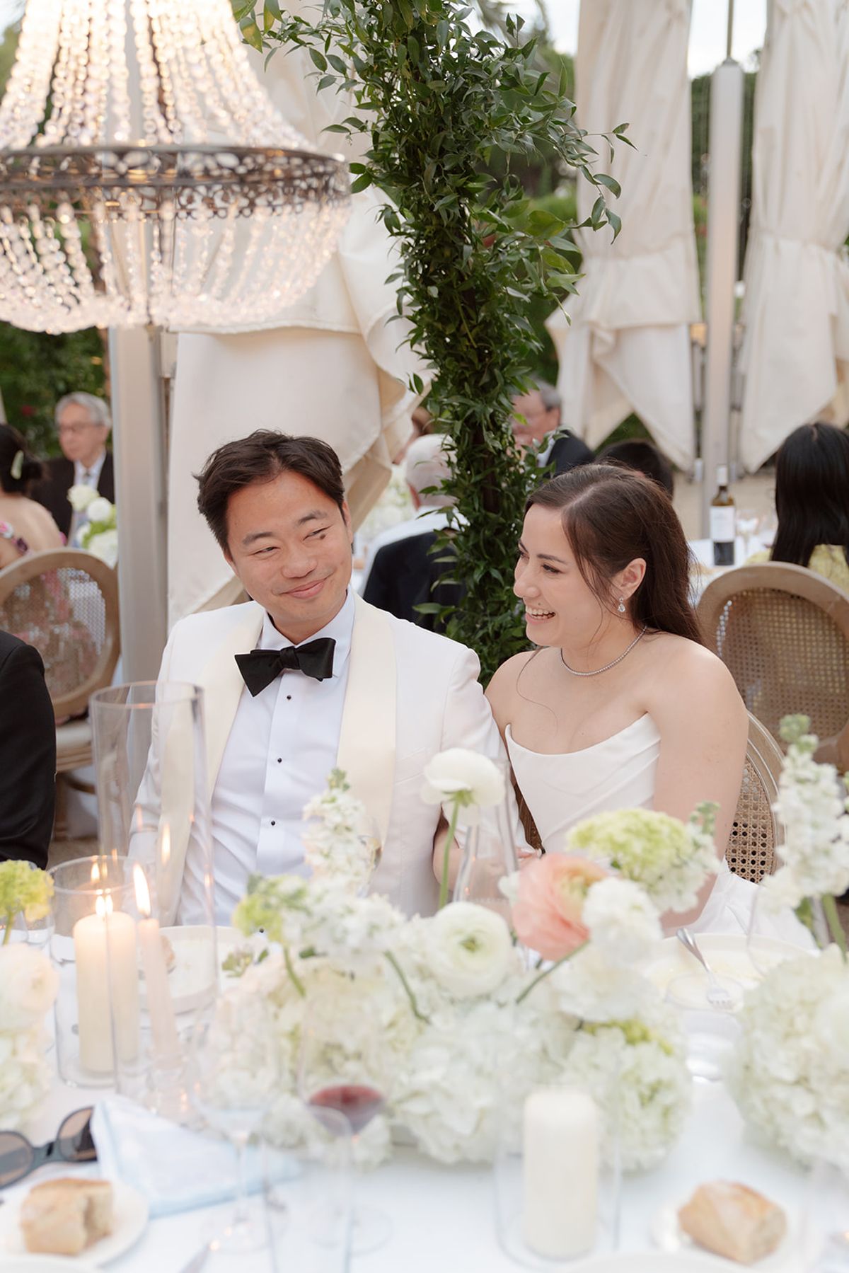 Stefan in a white tuxedo jacket and Carina smiling at the dinner table beneath a chandelier