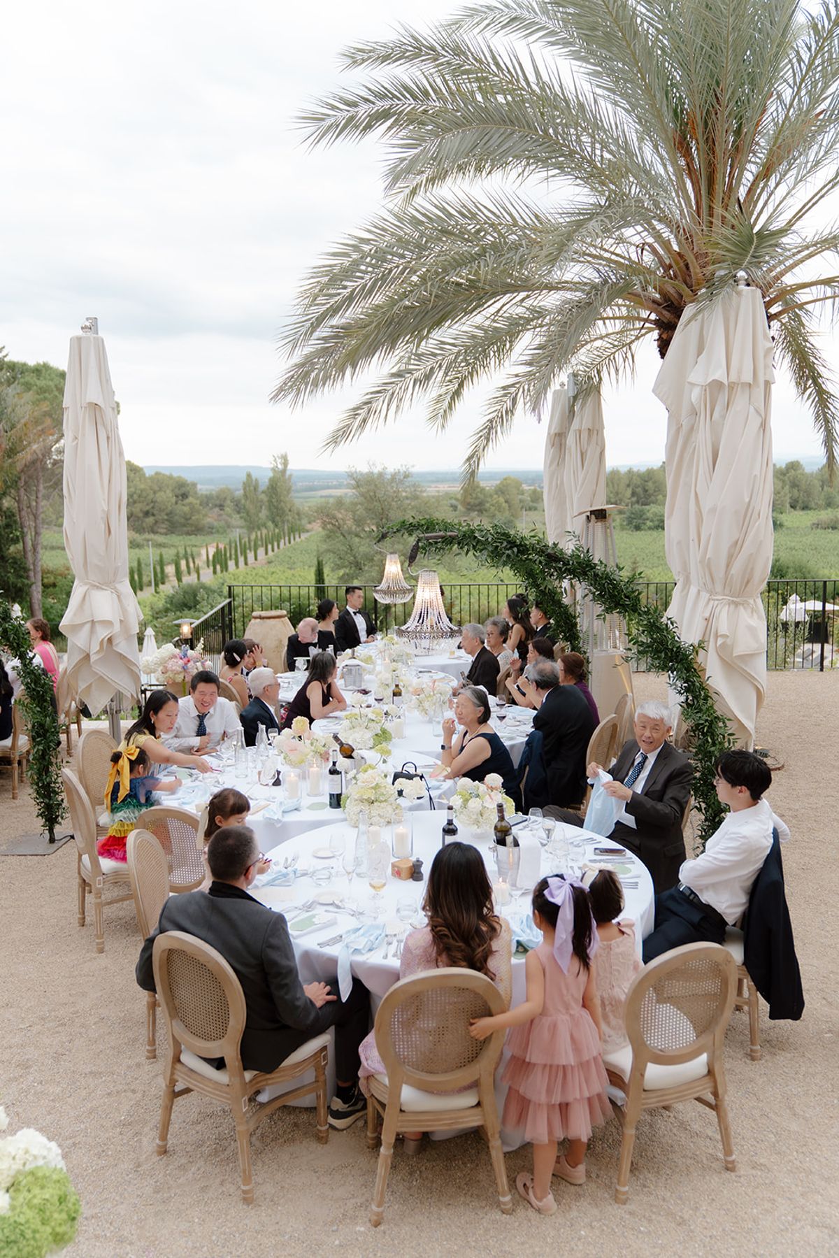 Guests seated at a round dinner table under a palm tree, a child in a pink dress beside them