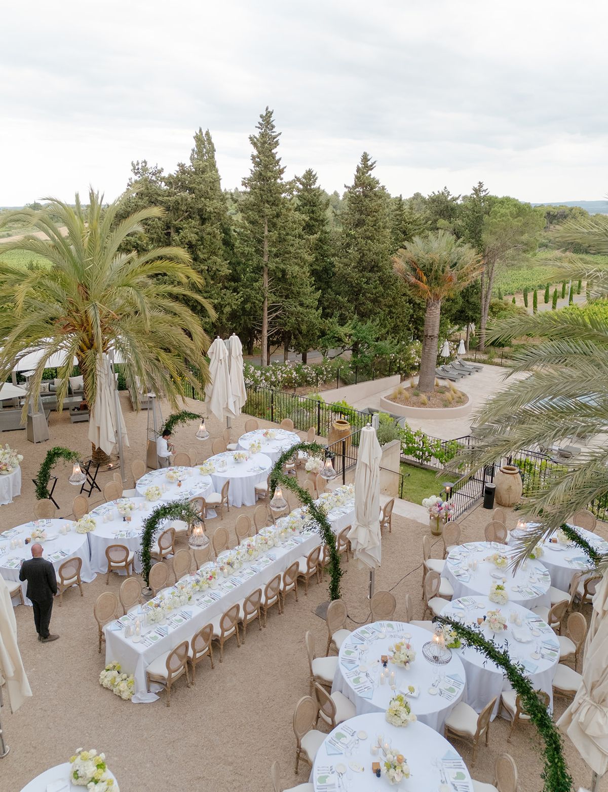 High view of the dinner terrace with long and round tables, green arches and palms