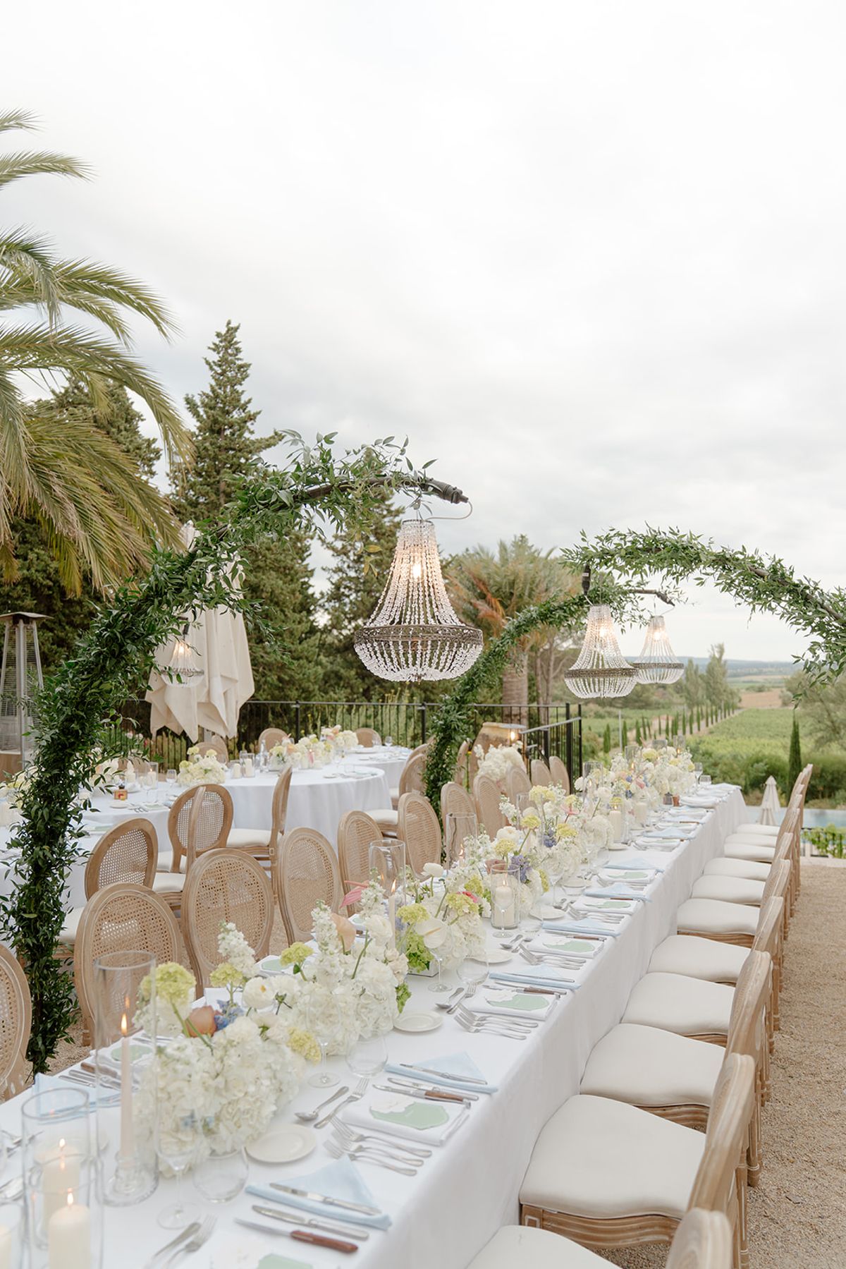 Long dinner table with low white florals running beneath green arches and chandeliers