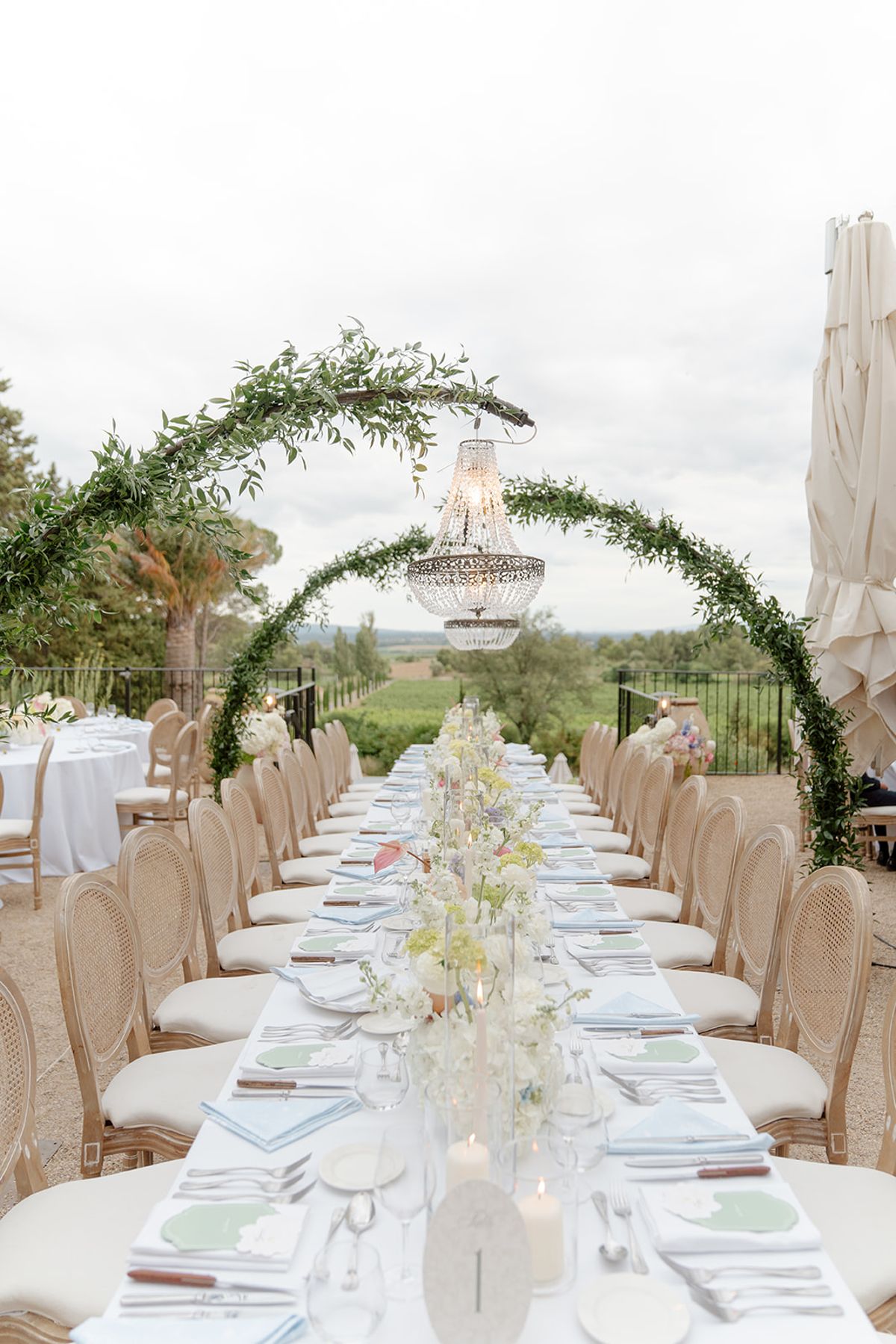 A long dinner table under green arches and crystal chandeliers, vineyards beyond