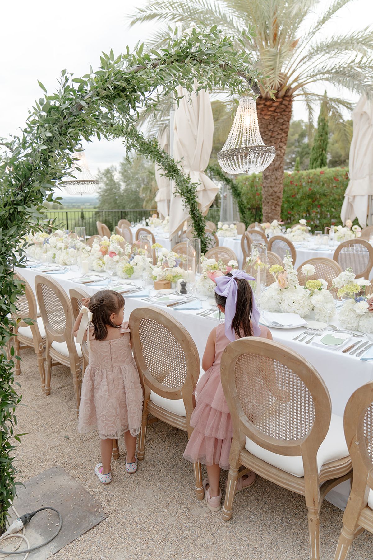 Two young guests in pink dresses looking at the dinner tables under a green arch and chandelier