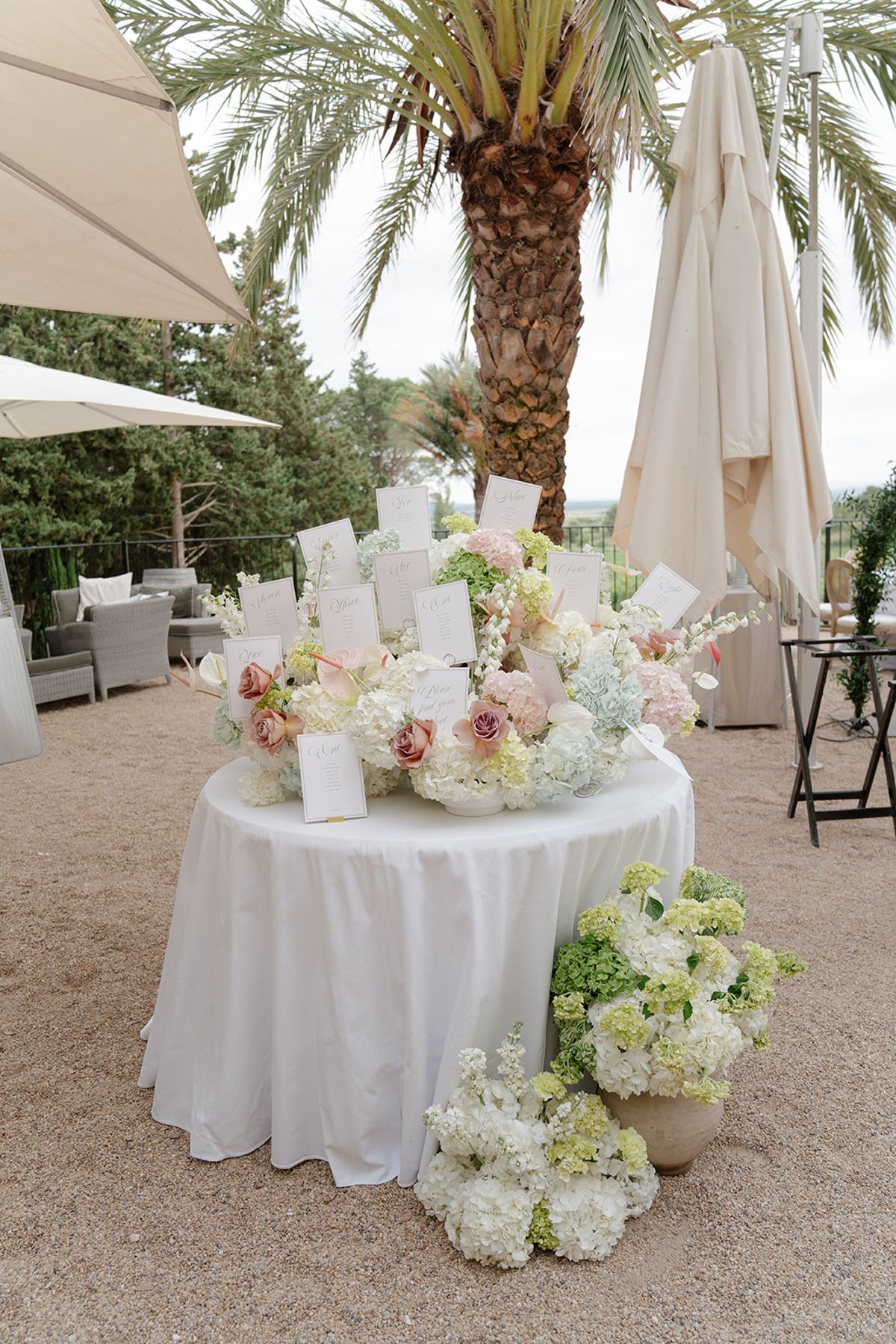 Escort card display of hydrangea and roses on a round table beneath a palm tree
