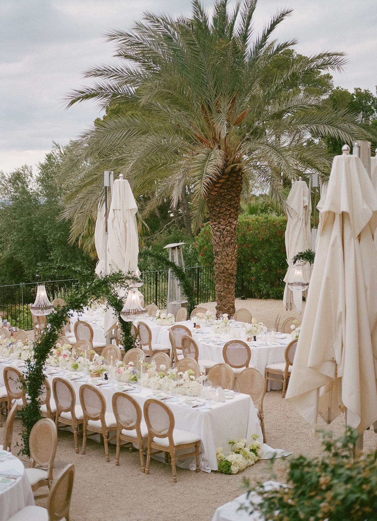 Dinner tables dressed in white beneath a palm tree, with parasols furled around them