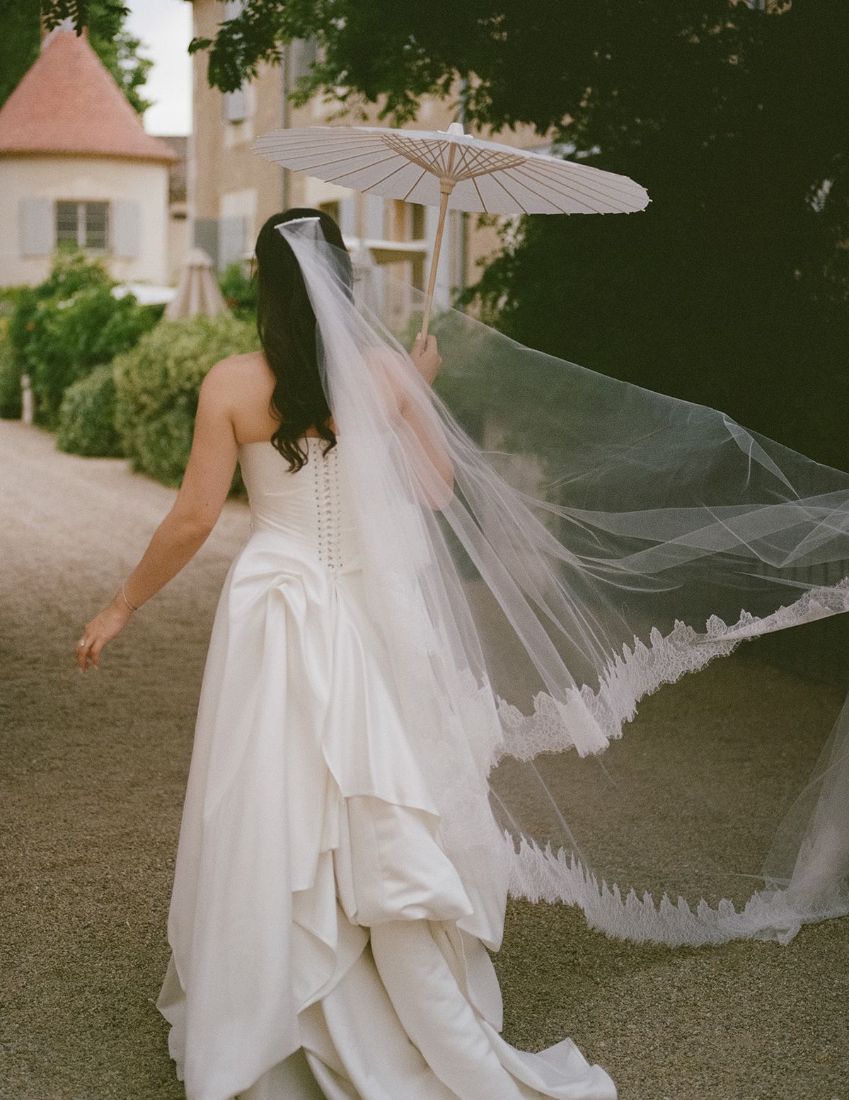 Carina seen from behind as her long lace-edged veil blows out on a gravel path